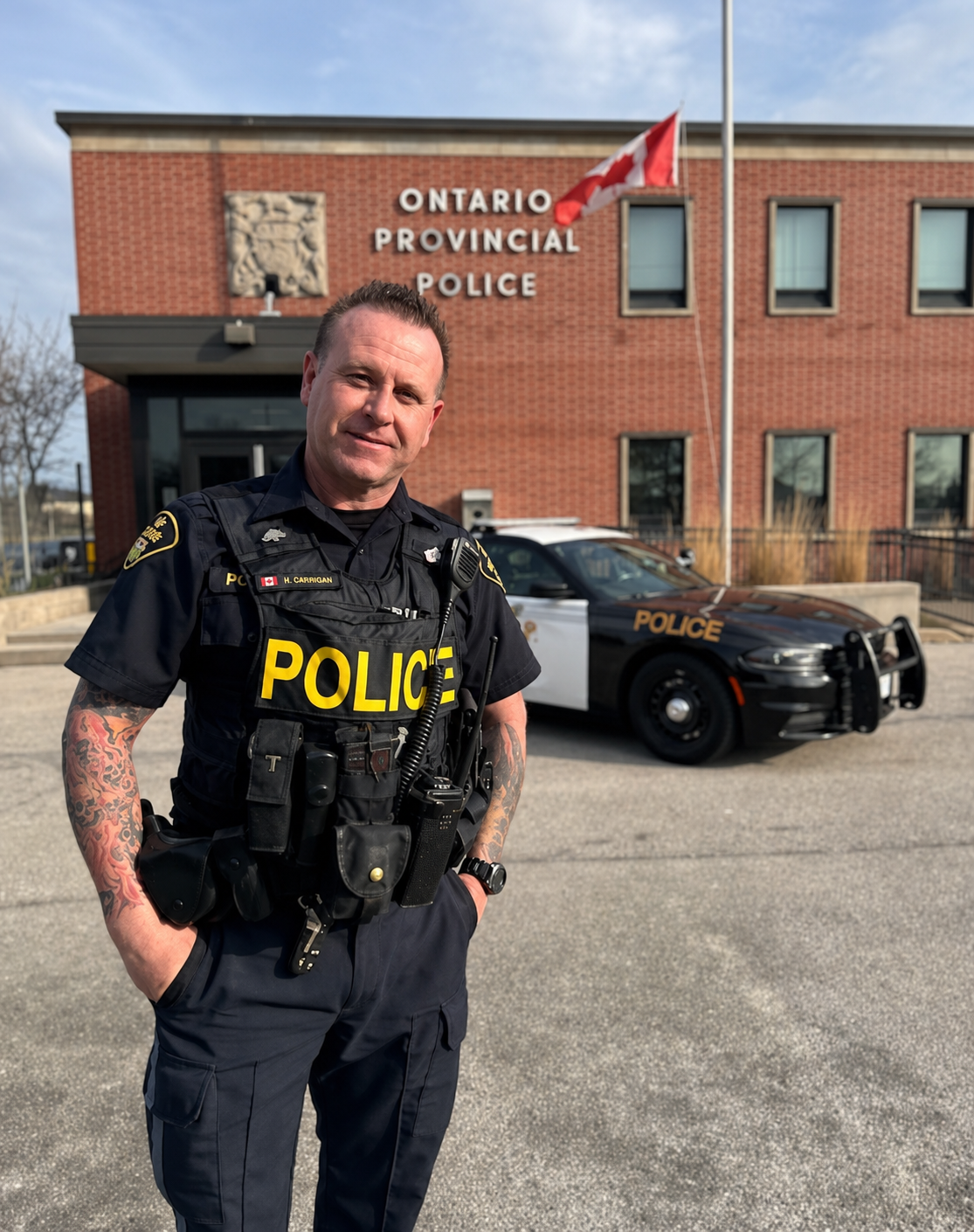 A police officer stands in front of the Ontario Provincial Police station, with patrol car parked behind him, wearing a black uniform with police gear, in daytime.