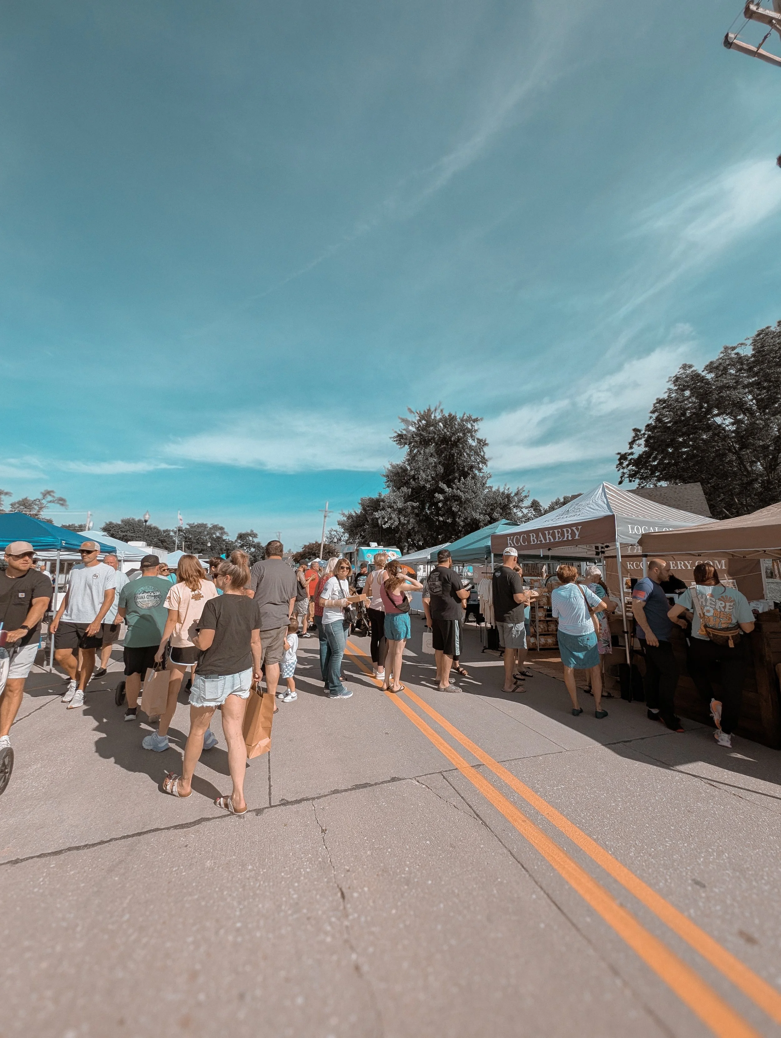 Crowd of people at an outdoor market under tents on a sunny day with a blue sky and trees.