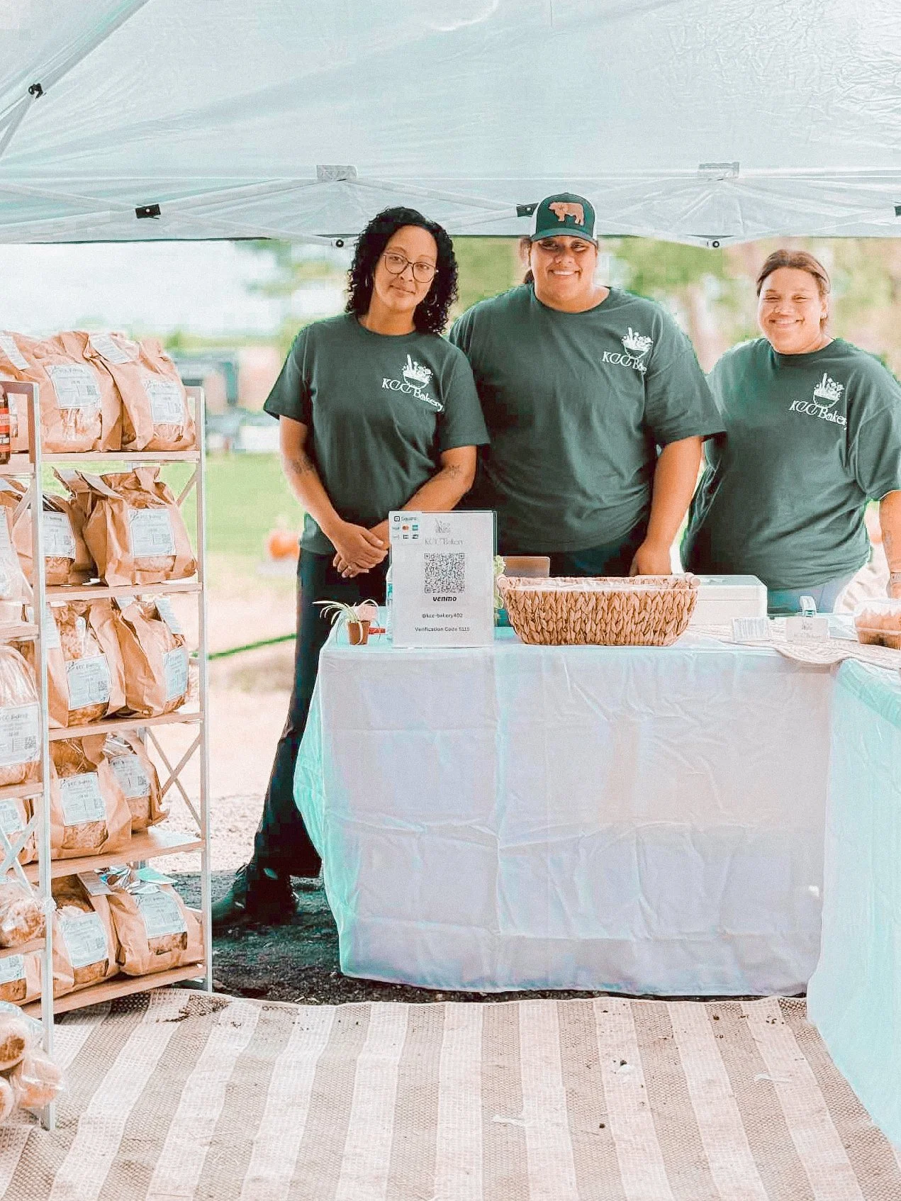 Three women standing behind a table at an outdoor market stall, with bags of baked goods on a shelf to the left and a QR code sign on the table, all wearing matching dark green t-shirts.