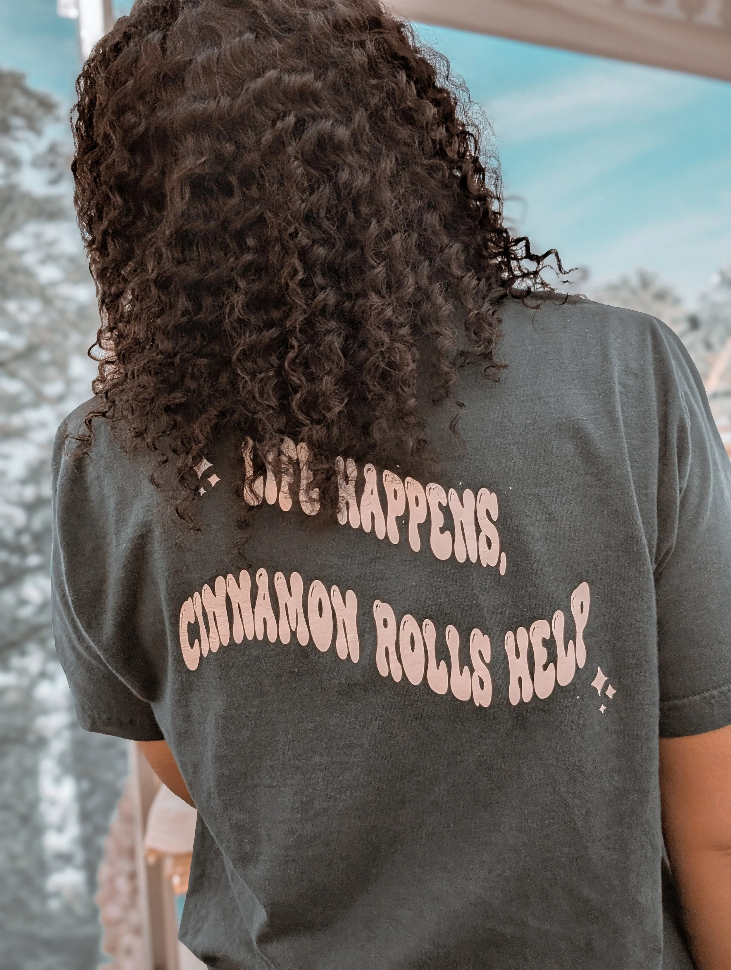 Back view of person with curly brown hair wearing a dark gray T-shirt with pink text, standing in an outdoor setting.