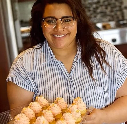 A woman with glasses and curly hair holding a tray of pink frosted cupcakes in a kitchen.
