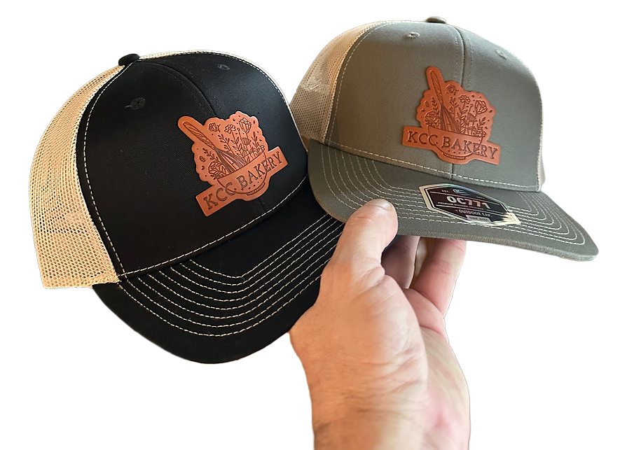 Two trucker hats with KCC Bakery patches clipped onto each other, one black and beige and one gray, against a black background.