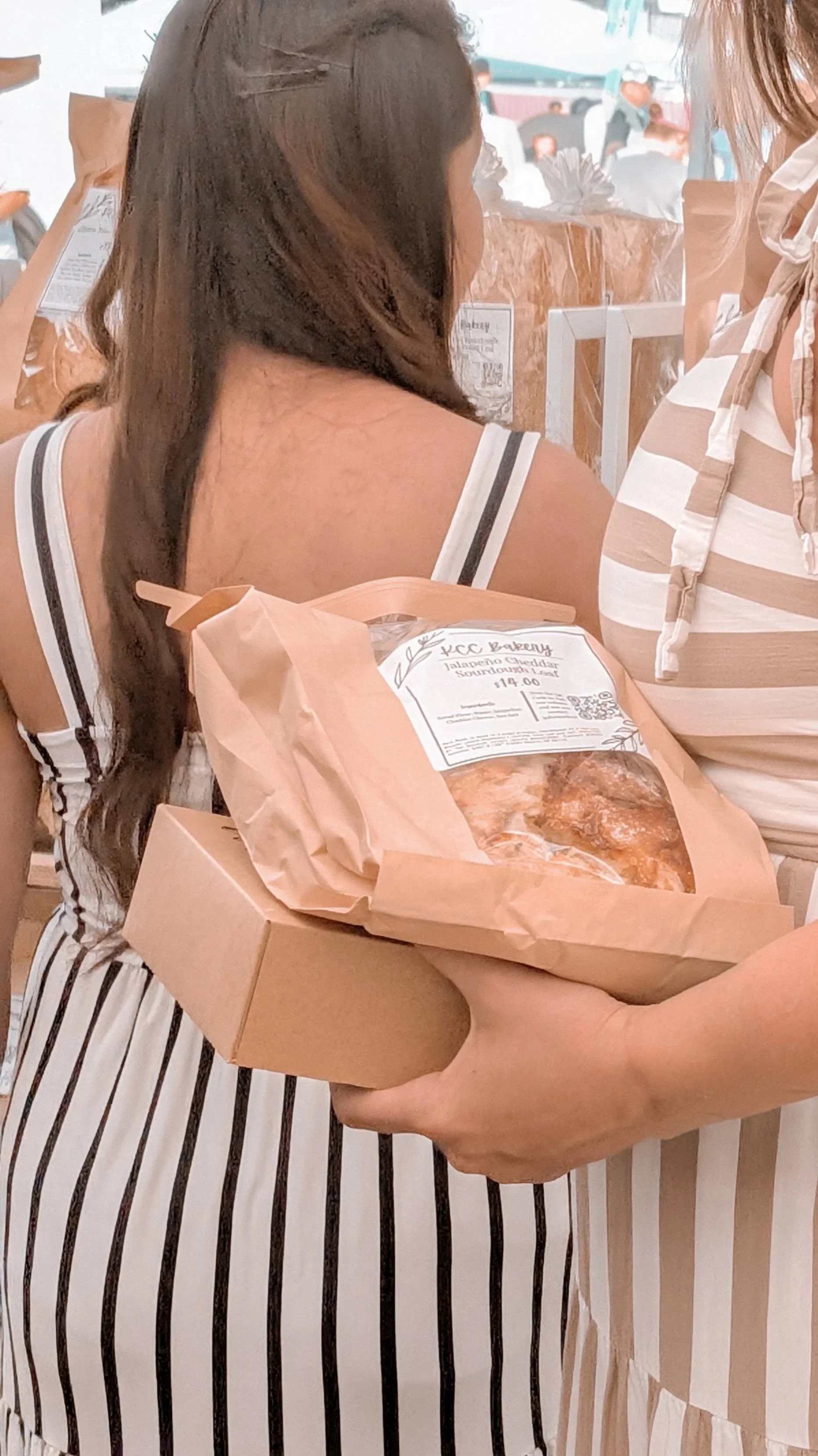 A person with long brown hair, wearing a sleeveless top, is receiving a box of baked goods at a bakery. The baked goods are labeled 'Jalapeño Cheddar Sour Cream Loaf' from KC Bakery, priced at $4.00.
