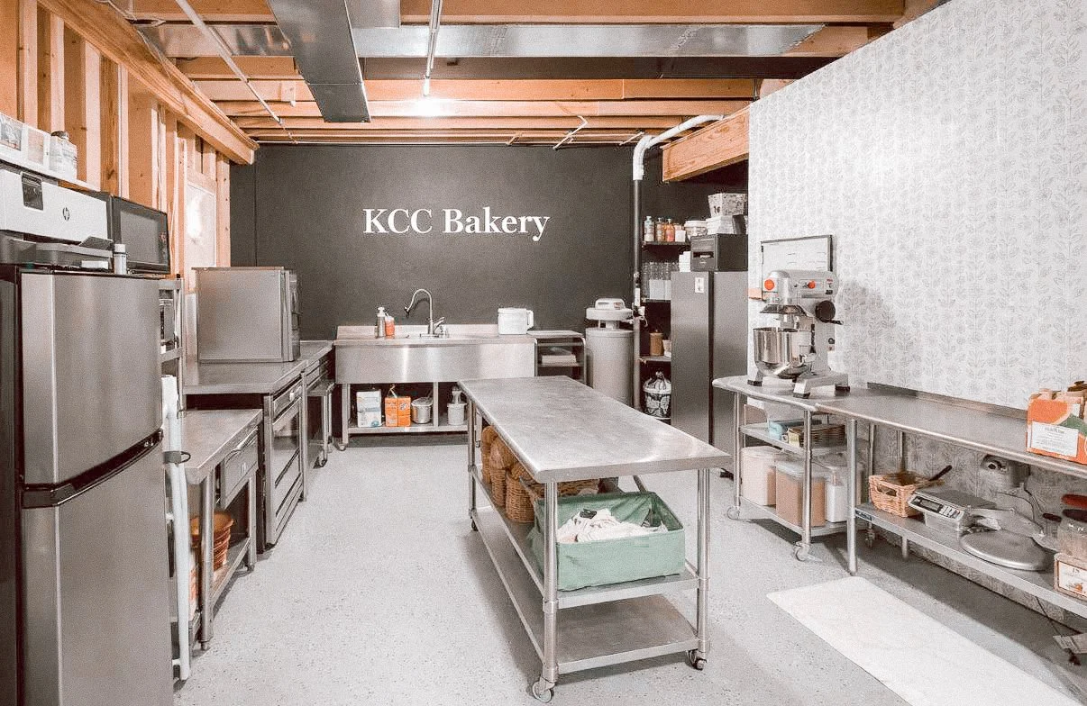 A commercial bakery kitchen with stainless steel appliances, tables, and shelves, and a black chalkboard wall with 'KCC Bakery' written on it.