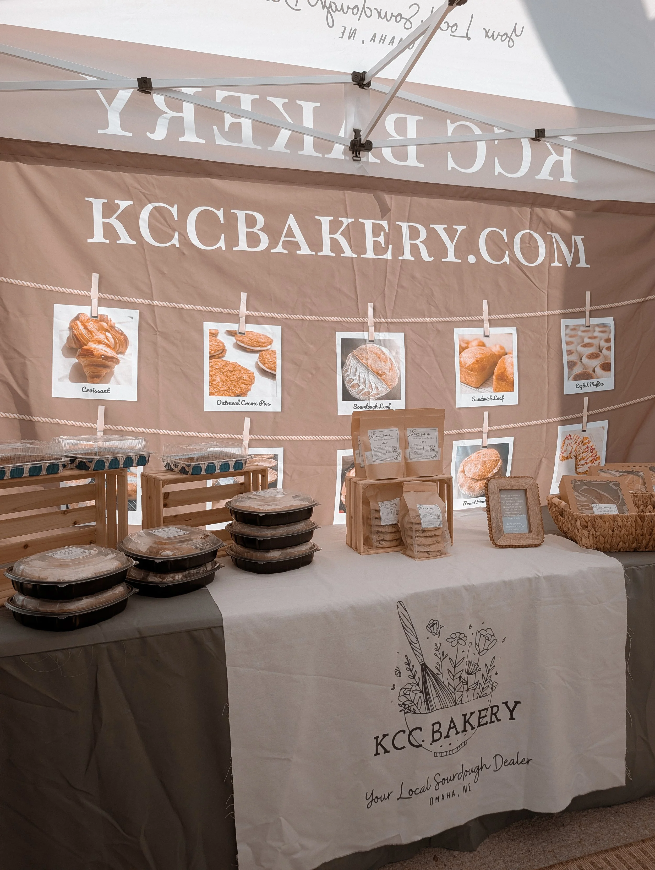 A bakery stall display with pre-packaged baked goods and a banner advertising KCC Bakery, including images of croissants, oatmeal creme pies, sourdough loaves, sandwich loaves, and English muffins.