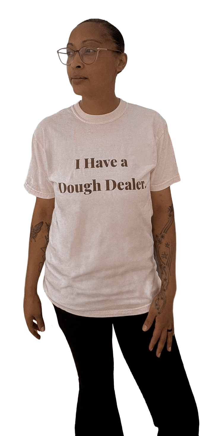 A woman with glasses, tattoos, and dark hair tied back, wearing a white T-shirt with the text 'I Have a Dough Dealer' and black pants.