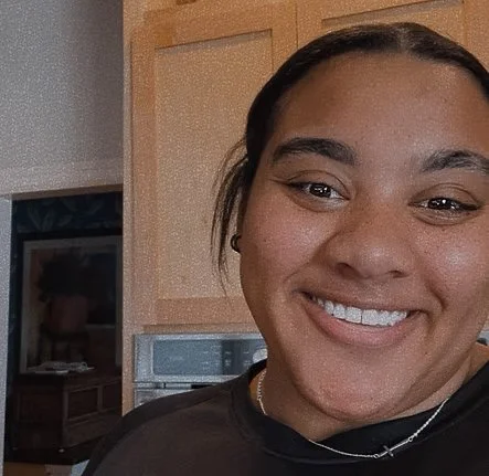 Close-up of a woman smiling in a home kitchen, with wooden cabinets and a microwave in the background.