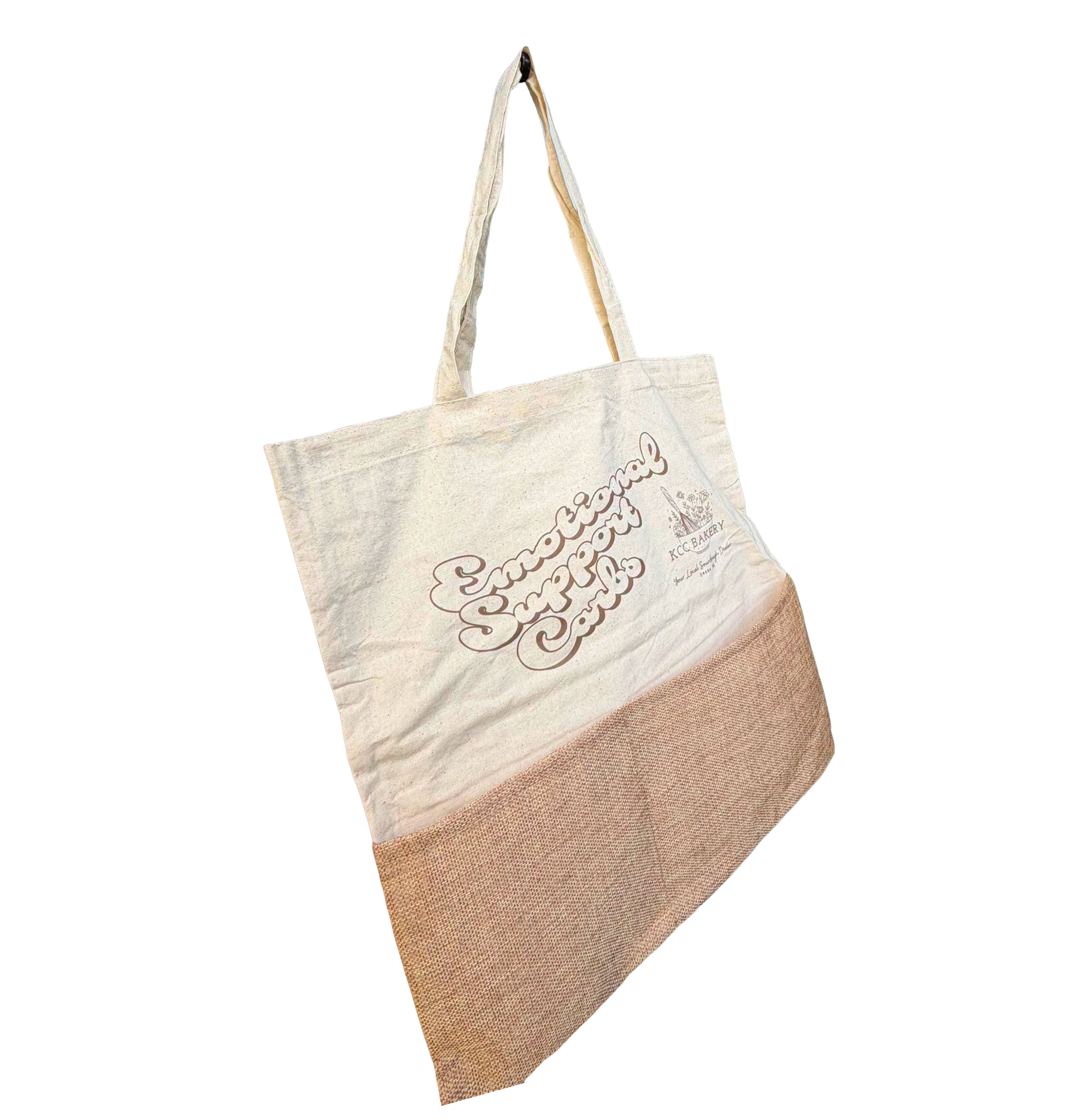 Close-up of a reusable beige canvas shopping bag with a brown logo that says 'Special Delivery' and 'KCC Bakery' on it, resting on a textured surface against a background with horizontal color bands.