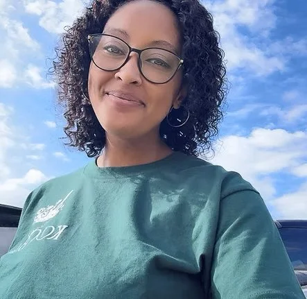 A woman with curly hair and glasses smiling outdoors against a blue sky with clouds, wearing a green shirt.