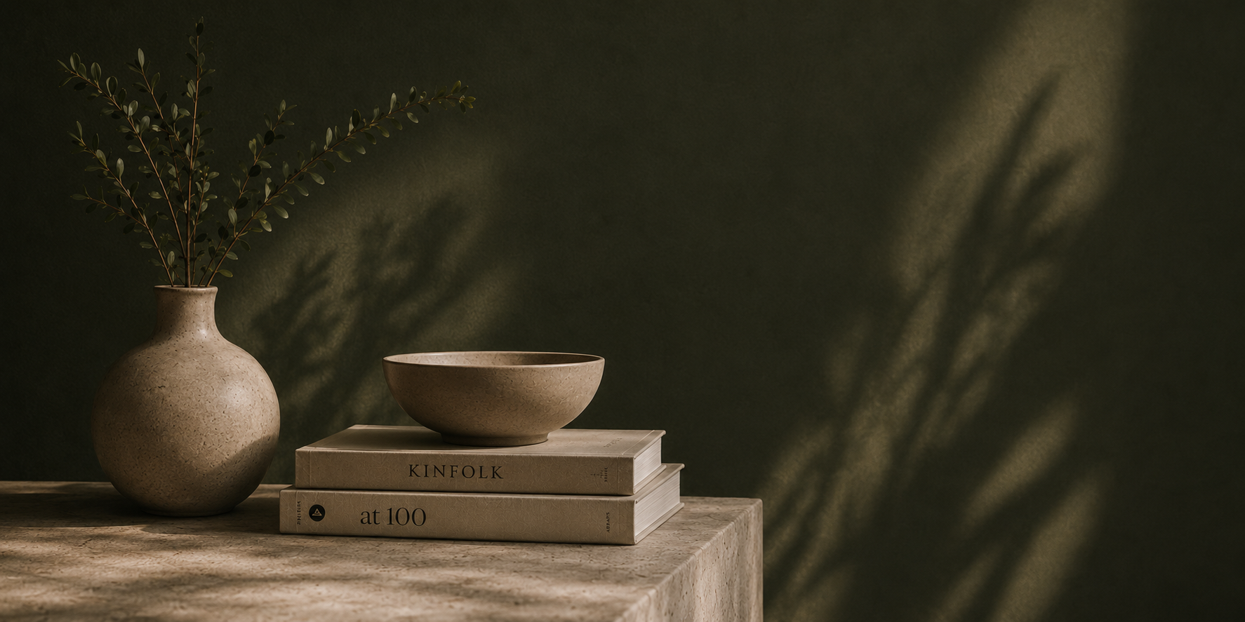 A beige ceramic vase with green leafy branches, a small beige ceramic bowl placed on top of two books titled 'KINFOLK' and 'at 100' on a textured beige stone surface, with a dark green wall background and shadows of leaves.