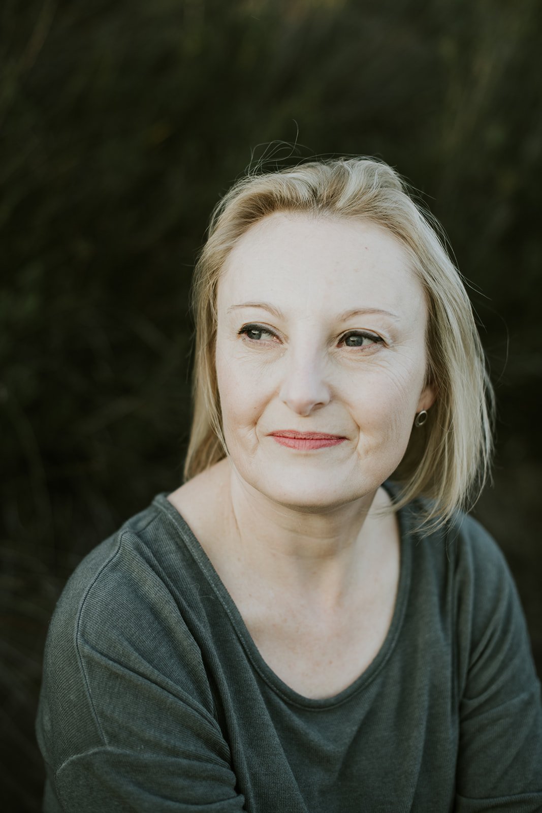 A woman with blonde hair and fair skin, wearing a dark gray top, sitting outdoors with a dark green, blurry background.