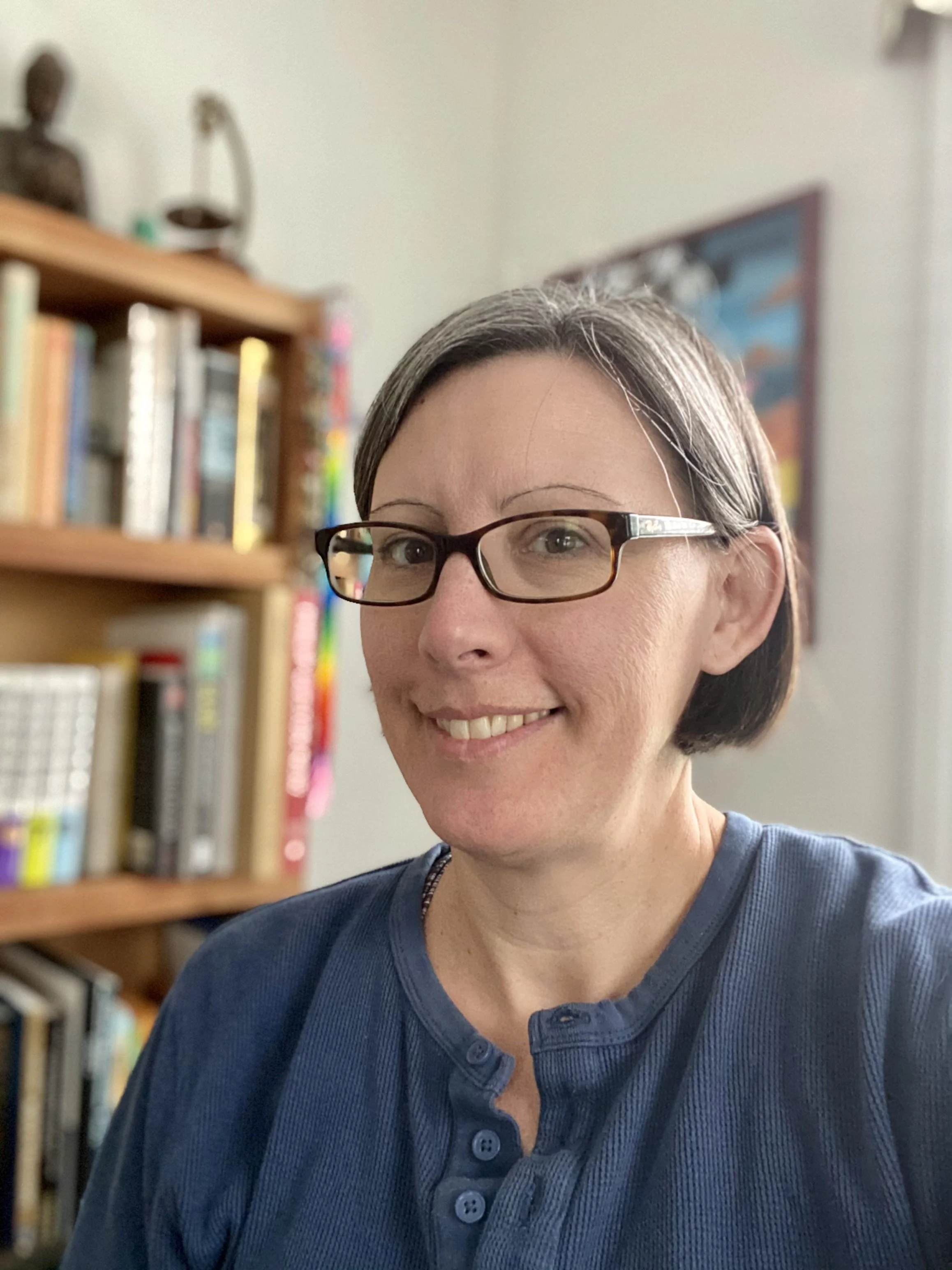 A woman with short hair and glasses smiling, wearing a blue shirt, standing in front of a bookshelf filled with books and a colorful poster in the background.