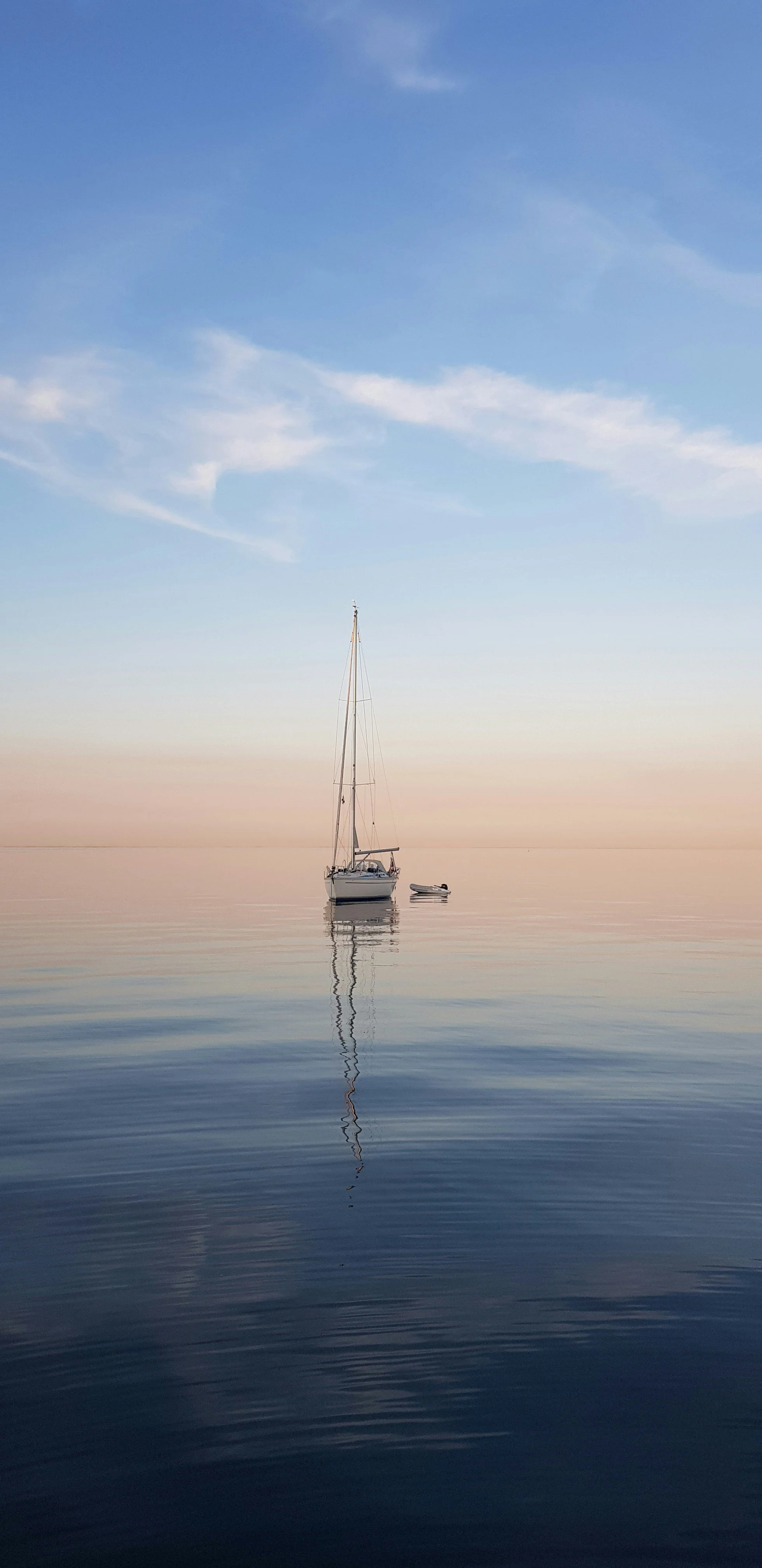 Sailboat anchored on calm water during sunrise or sunset, with a pastel-colored sky and soft ripples in the water.
