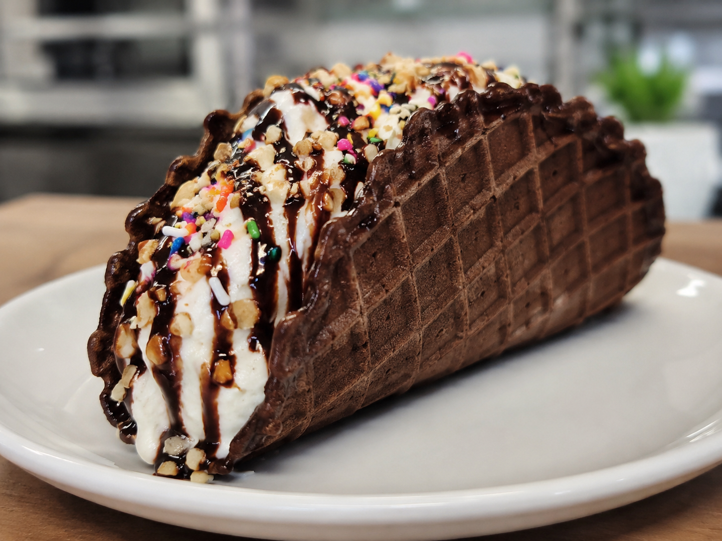Chocolate ice cream sundae in a waffle cone with chocolate syrup, sprinkles, and chopped nuts on a white plate.