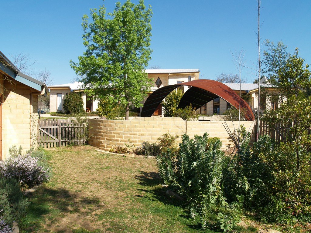A backyard with a large green tree, bushes, and a grassy lawn. In the background, there are residential houses with metal and brick structures. A curved metal sculpture or shelter is prominent in the middle of the scene, surrounded by a stone wall.