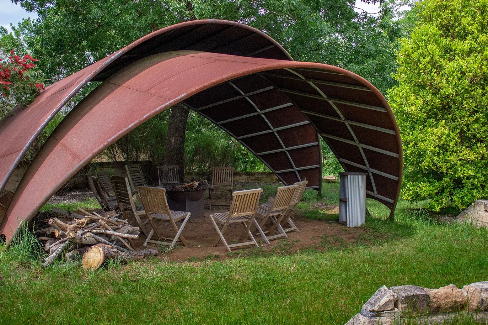 Outdoor wooden fire pit with around nine wooden chairs, a covered fire pit area, and a pile of firewood on grass, surrounded by trees and bushes.