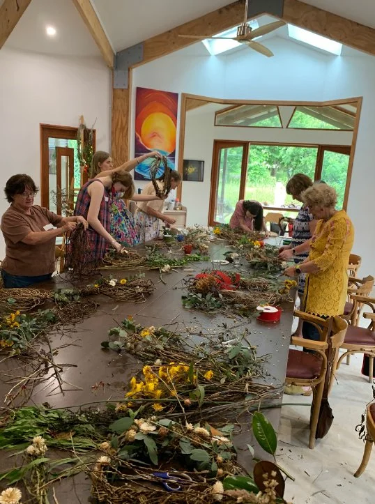 A group of women and girls making floral arrangements or wreaths around a large table in a bright craft room with large windows showing green trees outside.