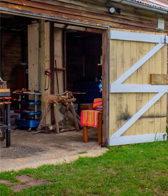 Open barn door revealing a workspace with woodworking tools, a wooden sculpture, and a colorful striped blanket on a chair, with a grassy area outside.