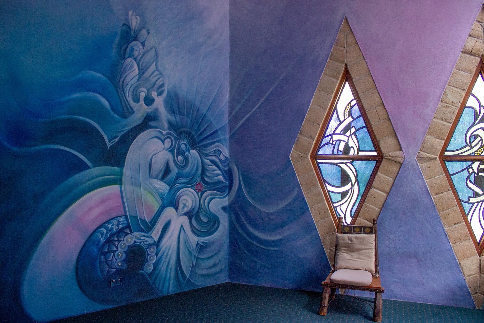 Interior of a room with a large, colorful mural on the left wall depicting abstract artistic figures and flowing shapes in blue and purple tones. The room has two tall, diamond-shaped stained glass windows with abstract patterns, framed by brick. There is an empty wooden chair with a cushioned seat placed near the windows.