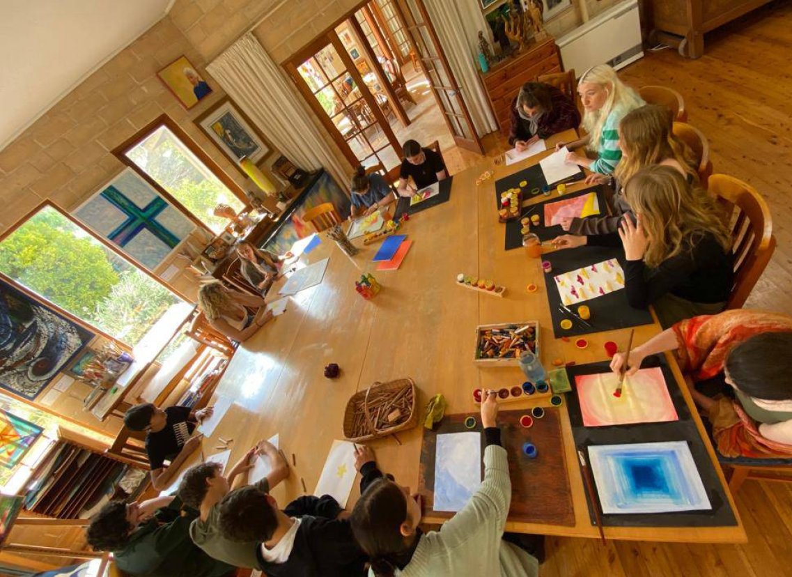 A group of people, including children and adults, gathered around a large wooden table engaged in painting and drawing activities in a well-lit room with large windows and artwork on the walls.