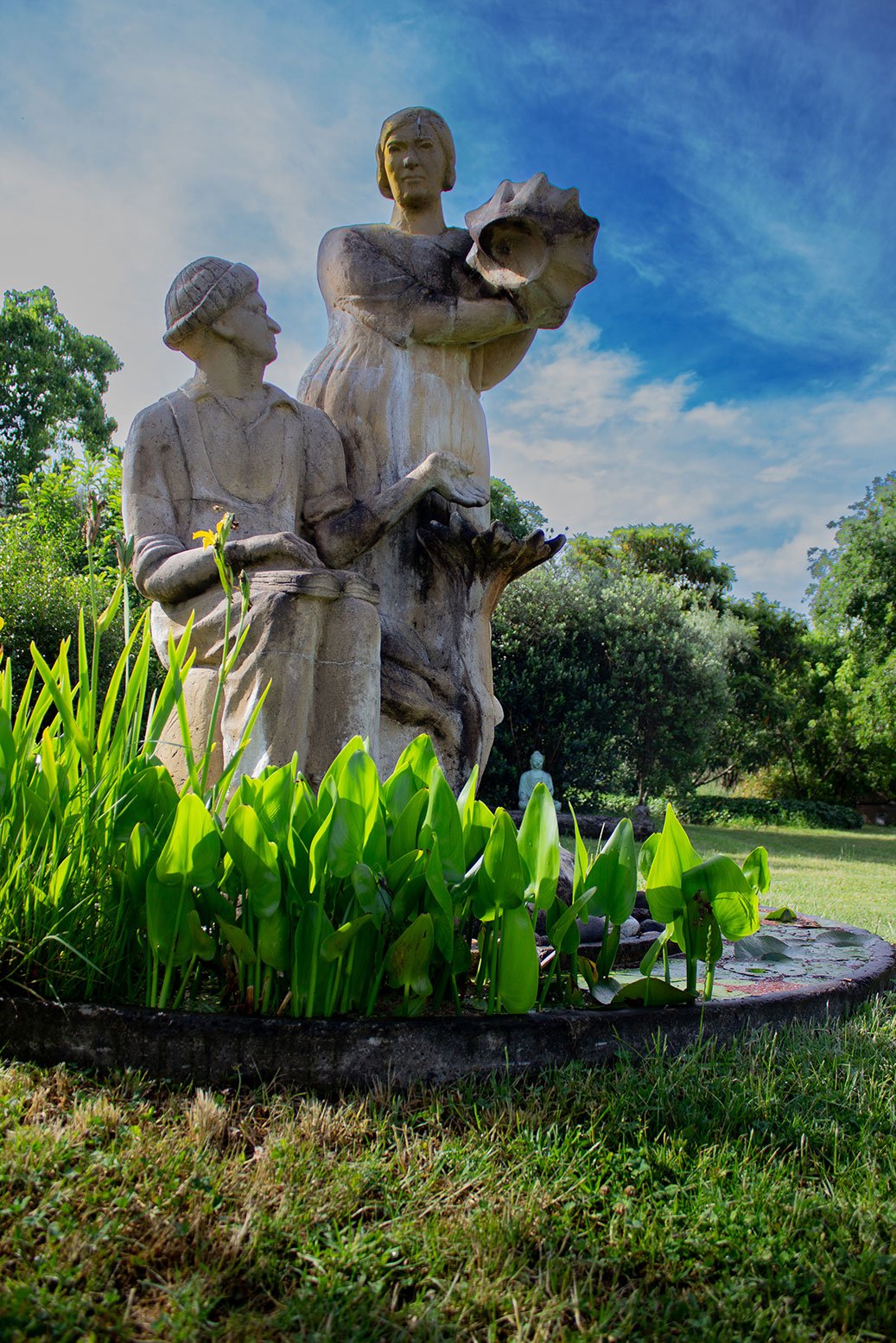 A stone sculpture of three people surrounded by green plants and trees under a blue sky.