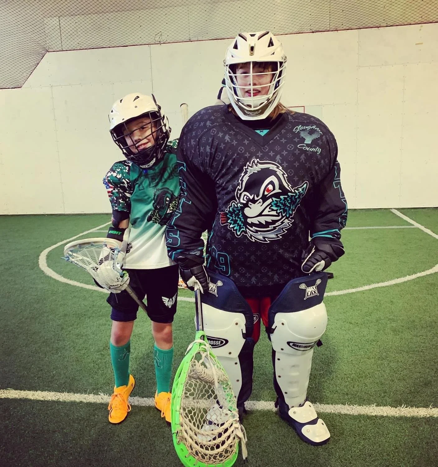 Two young box lacrosse players in protective gear inside an indoor rink.