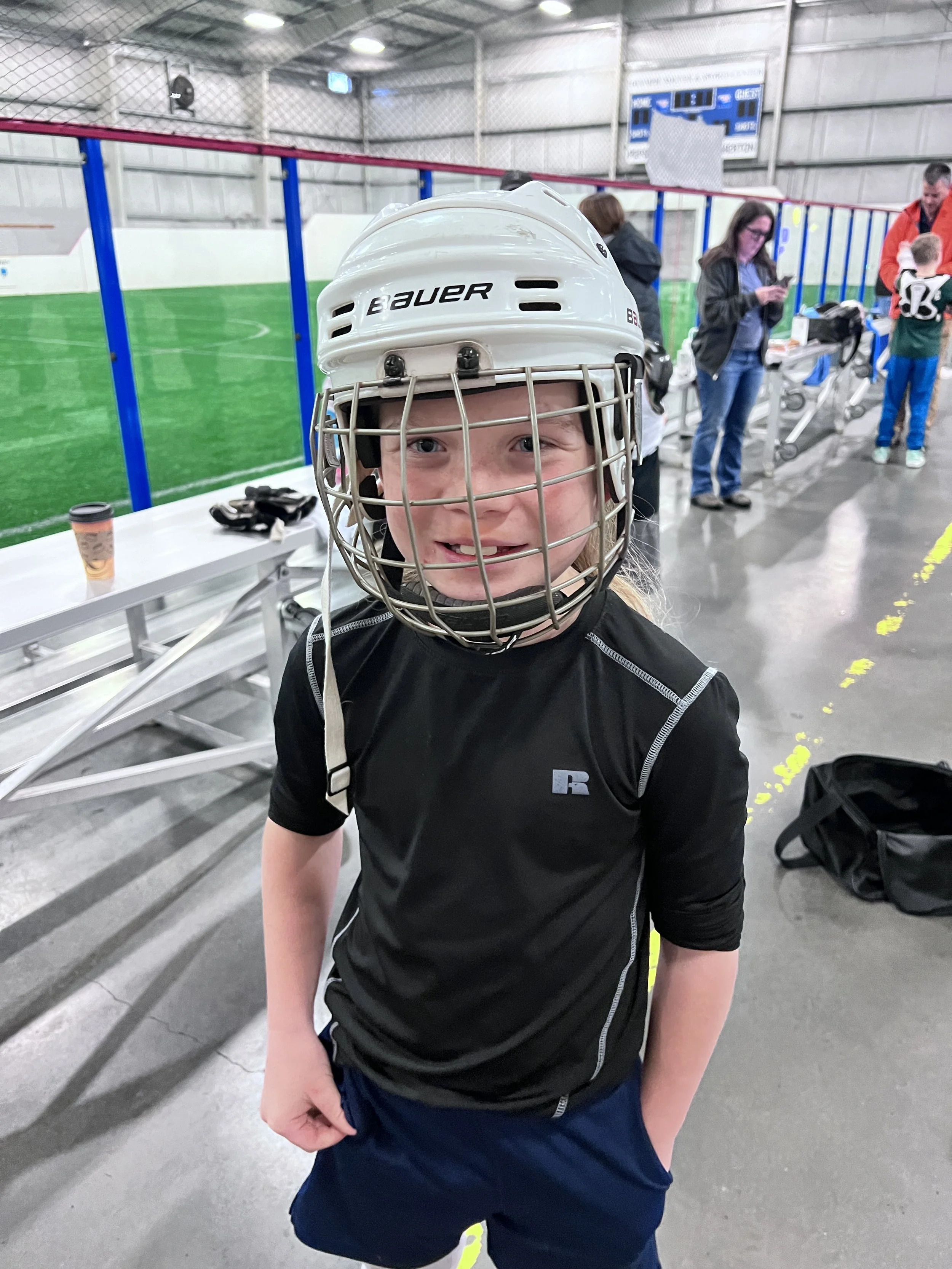 Young box lacrosse player wearing a white helmet inside an indoor rink.