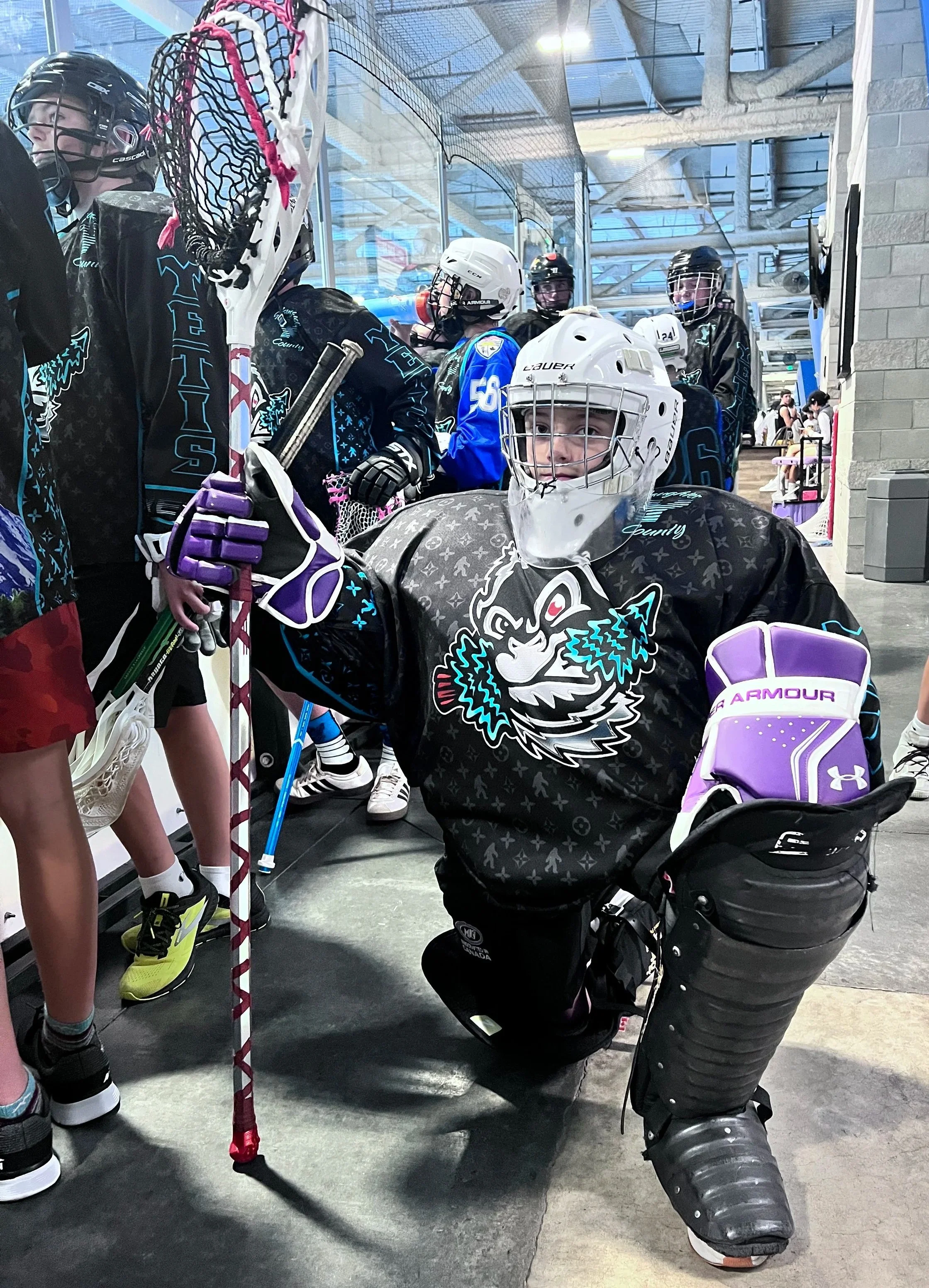 Youth box lacrosse players in helmets and team jerseys gathered inside an indoor rink.