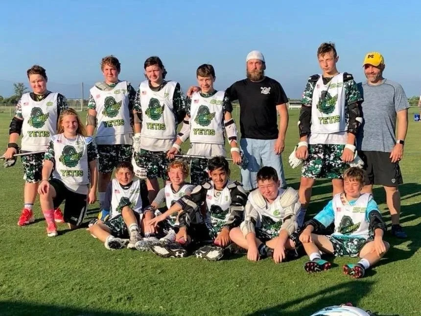 Youth lacrosse team and coaches posing together on an outdoor field.
