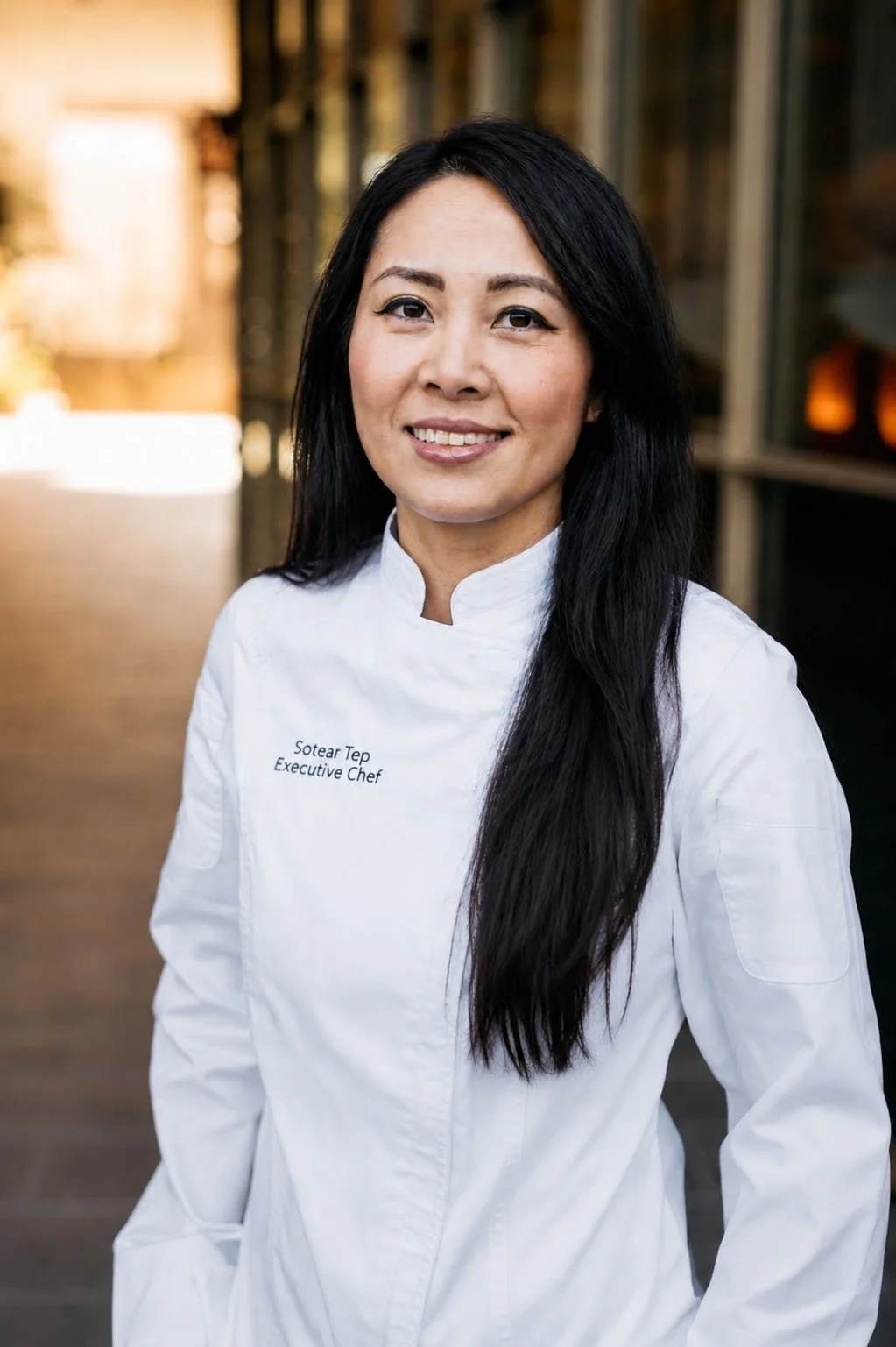 A woman chef in a white chef coat with the name and title "Sotear Tep, Executive Chef" embroidered on it. She has long black hair and is smiling, standing outdoors with blurred background.