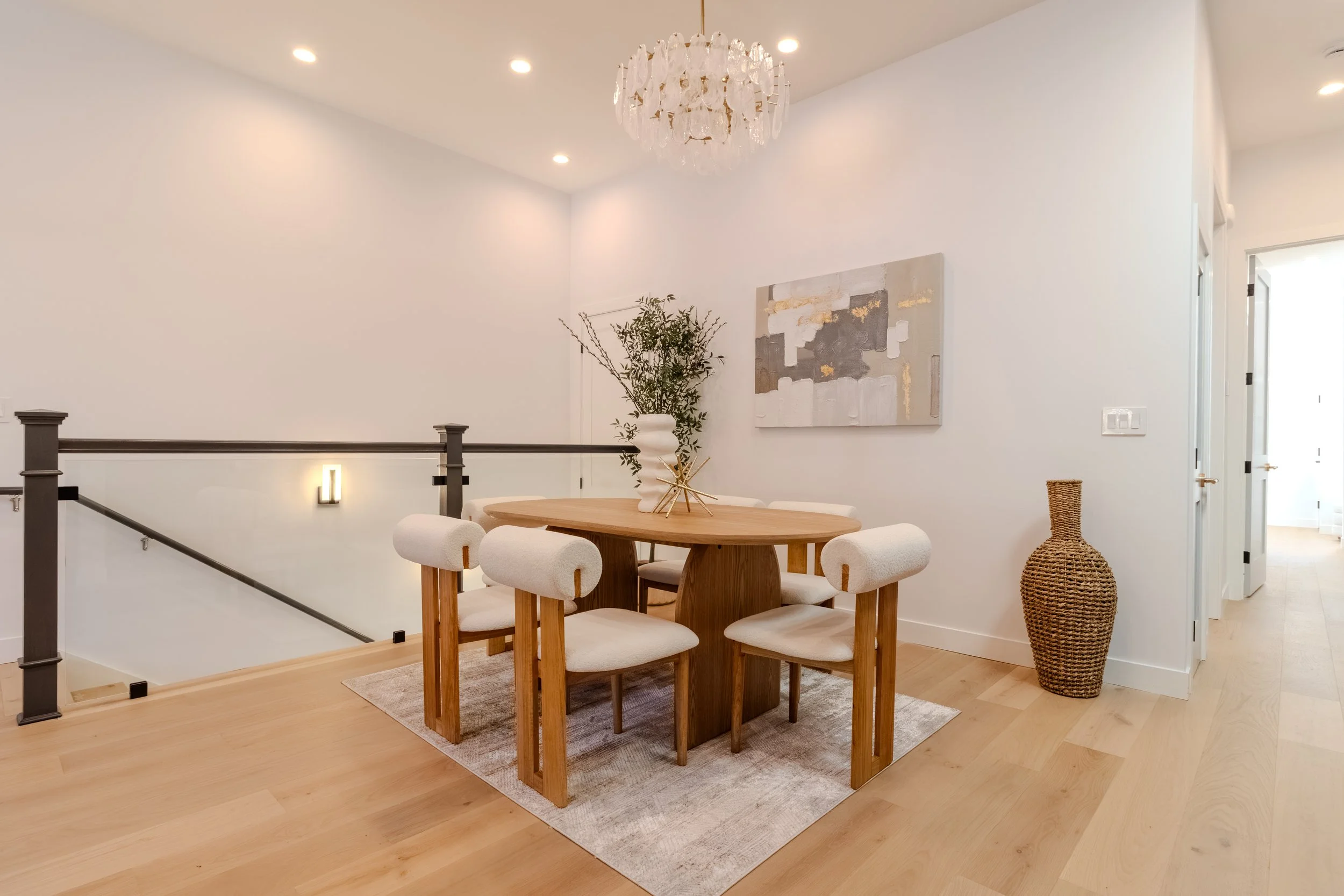 A dining area featuring a round wooden table with six upholstered white chairs, a large decorative vase with greenery, a framed abstract painting on the wall, a woven basket on the floor, and a chandelier hanging from the ceiling. The room has white walls, recessed lighting, and light hardwood flooring.