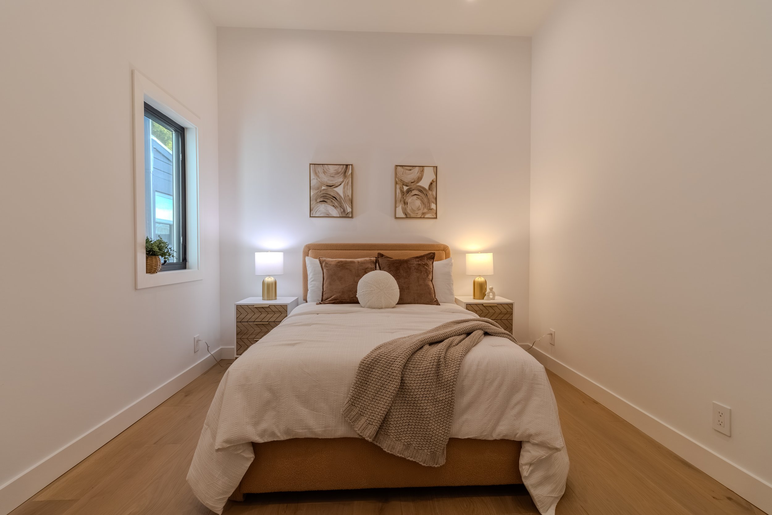 A cozy bedroom with a neatly made bed, beige headboard, and decorative pillows. There are two white nightstands with matching lamps on either side of the bed. Two framed abstract art pieces hang above the headboard. A window on the left side shows part of the exterior with a plant on the windowsill.