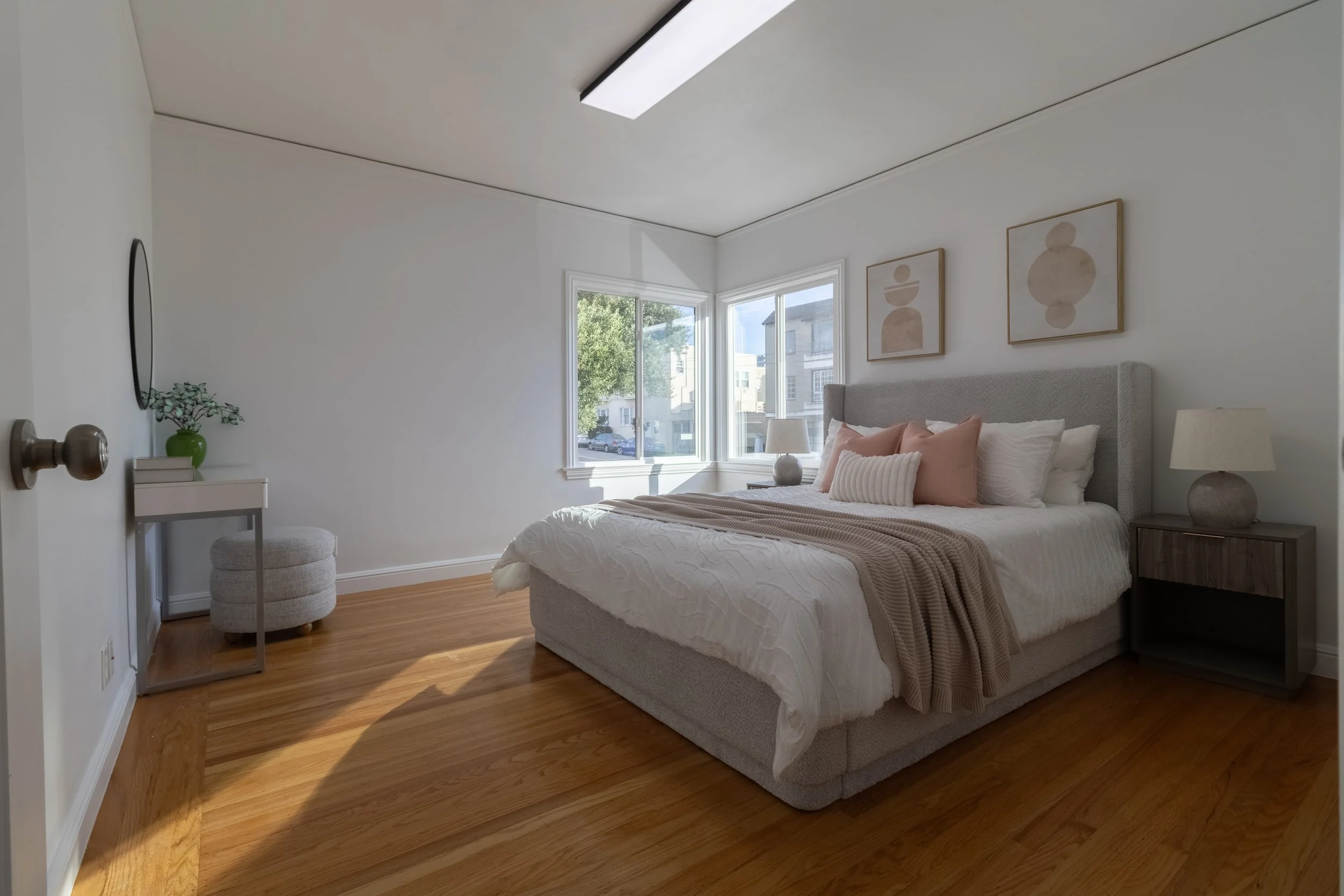 A bedroom with white walls and a wooden floor, featuring a large bed with white, beige, and pink pillows. There is a bedside table with a lamp and a small green vase with plants on a small white table, along with other framed artwork on the wall.