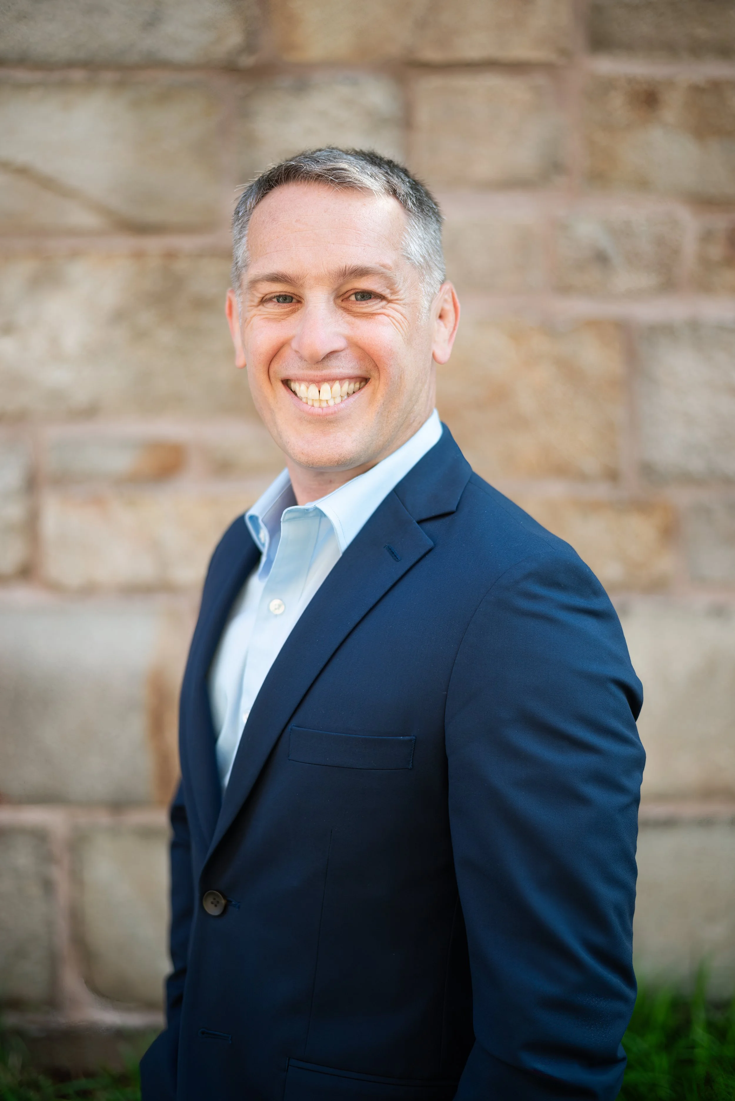 A smiling man wearing a navy blazer and white shirt, standing in front of a brick wall.