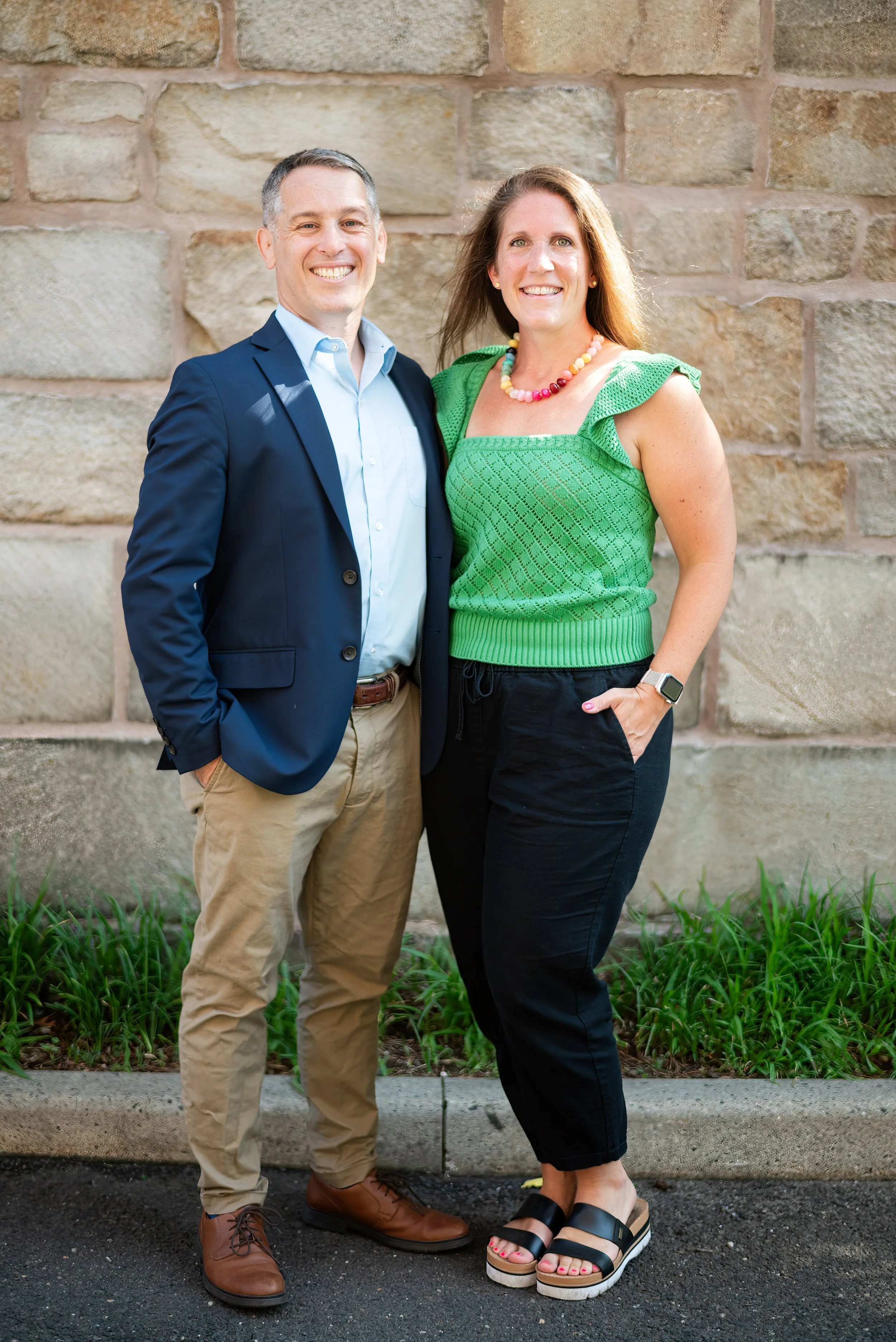 A man in a dark blue blazer, light blue shirt, and khaki pants standing next to a woman in a green top and black pants, both smiling, outdoors in front of a brick wall with grass in the foreground.