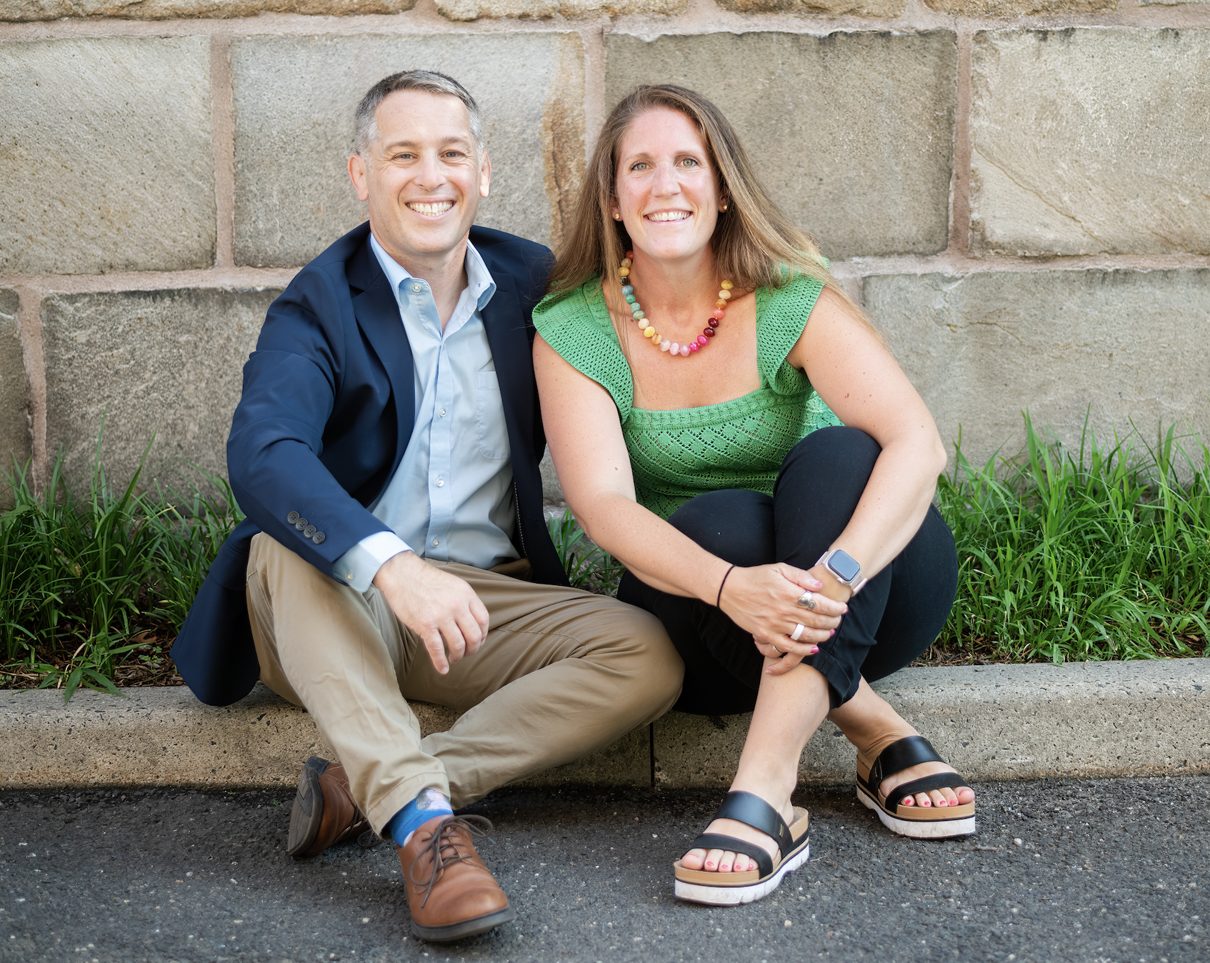 A man and a woman sitting on a concrete ledge in front of a brick wall, smiling at the camera. The man is dressed in a navy blazer, light blue shirt, khaki pants, and brown shoes. The woman is wearing a green sleeveless top, black pants, and black platform sandals, with a colorful beaded necklace and a smartwatch.
