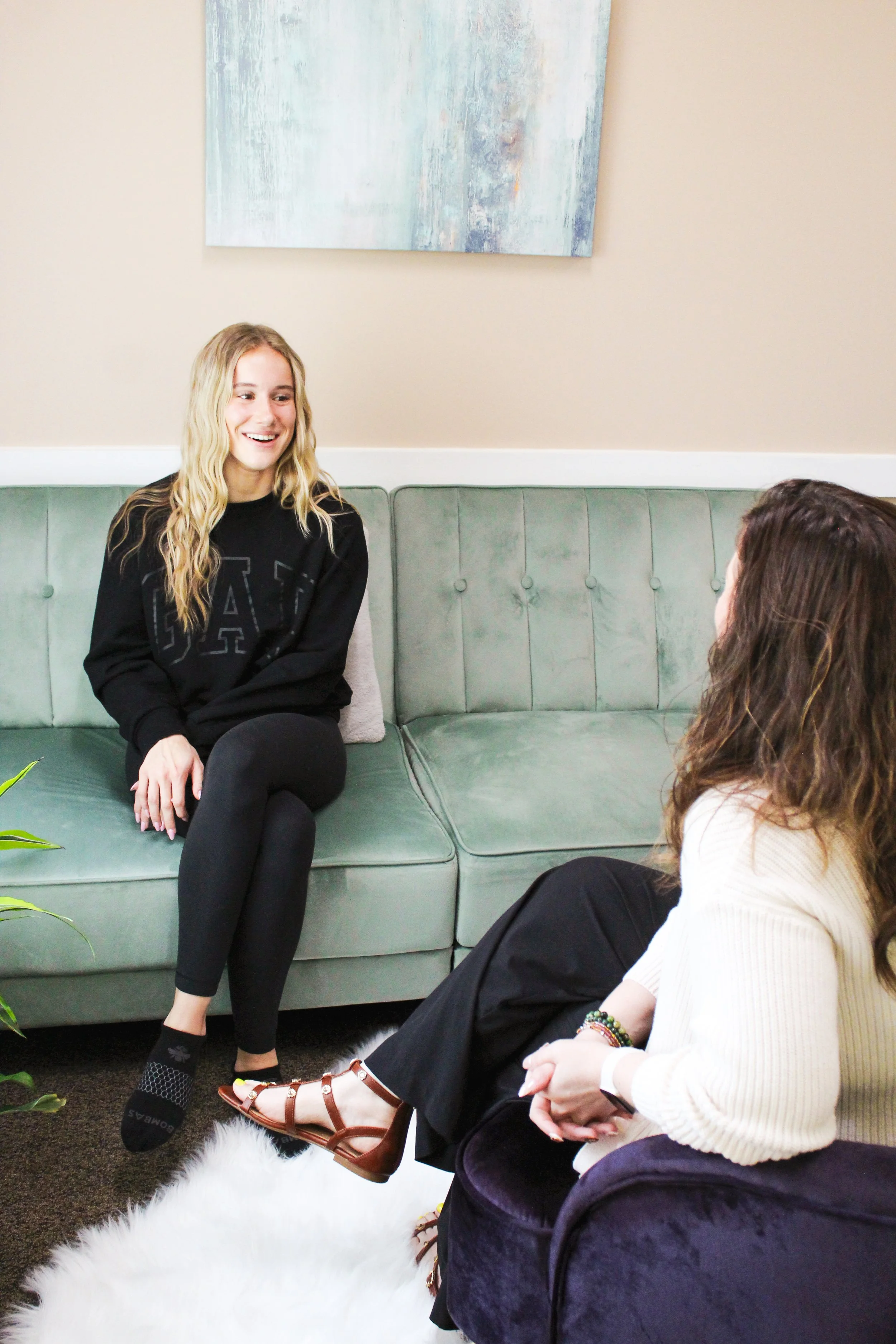 Two women sitting on a green sofa, engaged in conversation. One woman with long blonde hair and black clothing is smiling. The other woman has wavy brown hair, wears a white sweater, black pants, brown sandals, and has colorful bracelets. There is a white fluffy rug on the floor, and a large abstract painting hangs on the beige wall behind them.