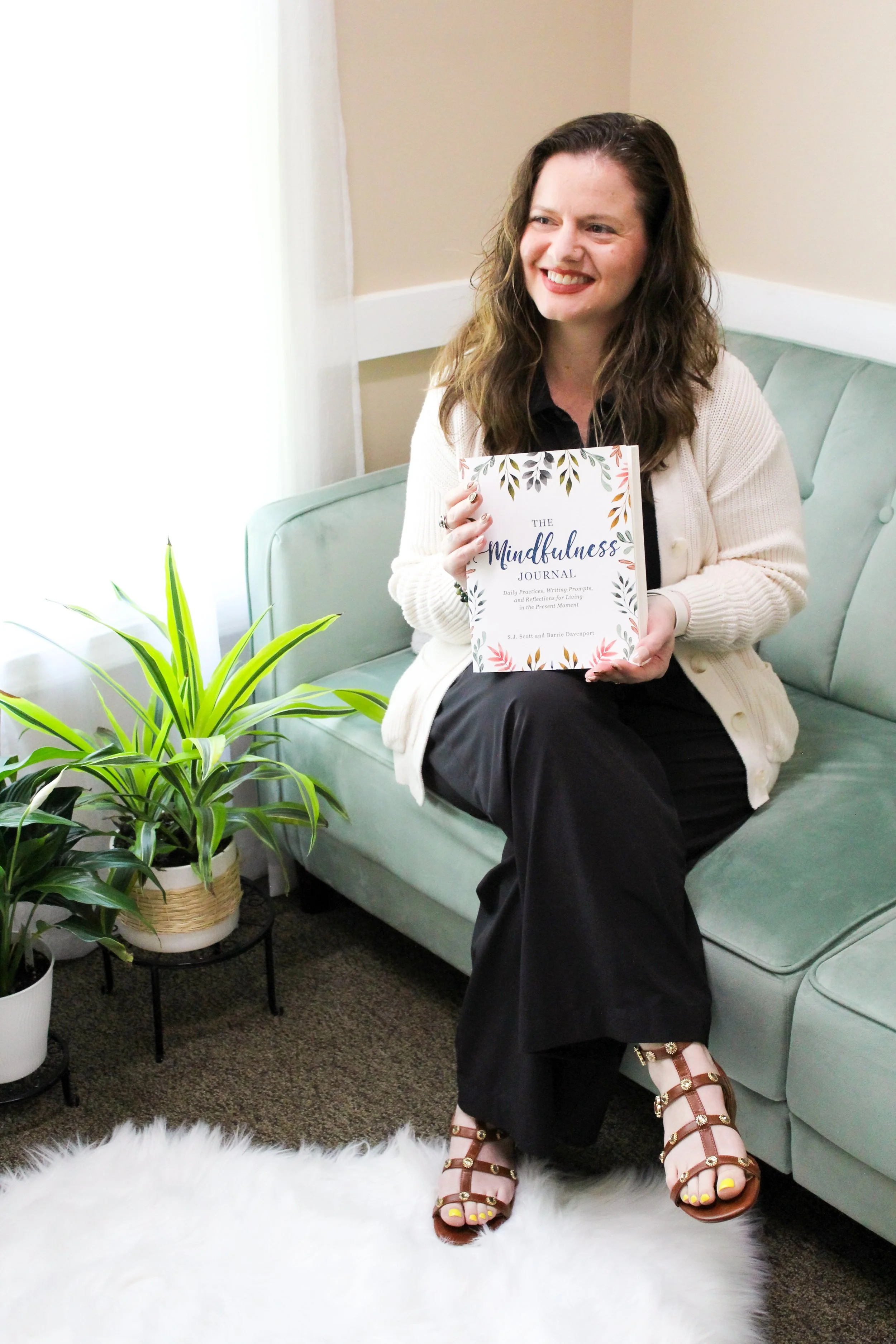A woman with long, wavy brown hair sitting on a light green sofa, smiling and holding a book titled 'The Mindfulness Journal'; she is wearing a black top, a white cardigan, black pants, and strappy brown sandals with gold embellishments; potted plants are nearby, and a white fluffy rug covers the floor in front of her.