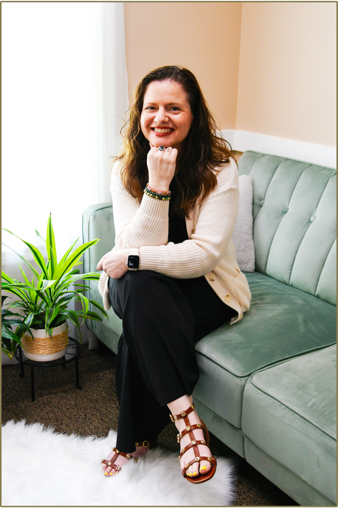 A woman sitting on a green velvet sofa, smiling and resting her chin on her hand, wearing a beige sweater, black pants, and brown gladiator sandals with gold embellishments, in a cozy living room with a potted plant, white fluffy rug, and soft natural light from a window.