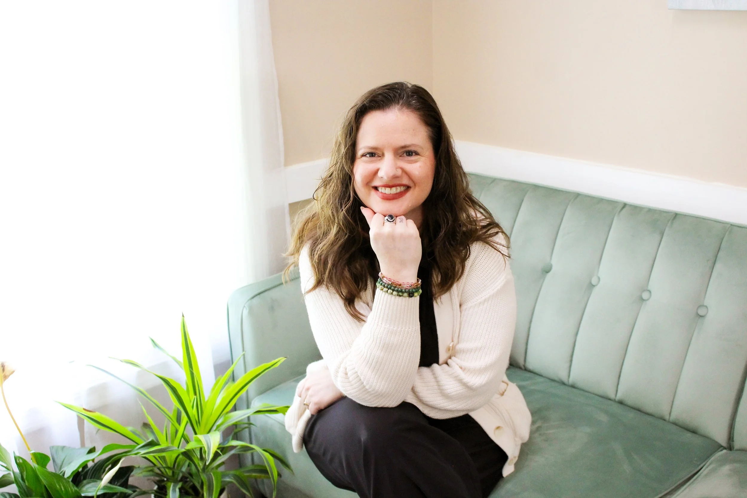 A woman with long, wavy brown hair and a friendly smile is sitting on a light green tufted bench in a room with beige walls and natural light. She is wearing a cream-colored sweater, black pants, multiple colorful bracelets, and a ring on her finger. She rests her chin on her hand and is near a potted green plant with long, pointed leaves.