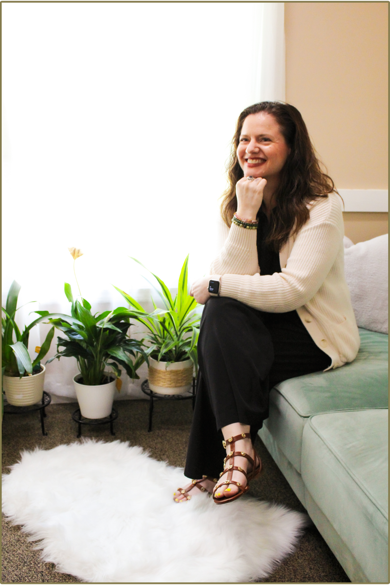 A woman sitting on a green sofa next to a fluffy white rug. She has long dark hair, is smiling, and resting her chin on her hand. She is dressed in a cream-colored cardigan, black pants, and strappy brown sandals. There are several potted green plants on black plant stands behind her. The background features a beige wall, a window with white curtains, and a white shaggy rug on a brown carpet.