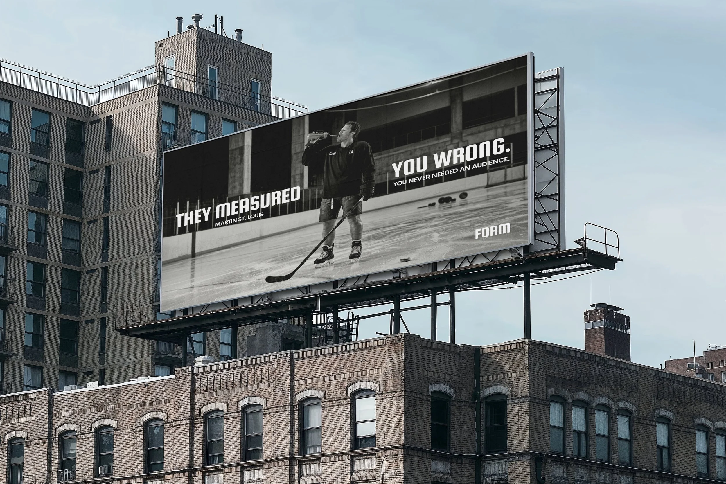 A large outdoor billboard on top of a building displays a black-and-white image of a man with hockey skates drinking from a water bottle while standing on a hockey rink with hockey sticks lying on the ice. The text on the billboard reads: "They measured. You wrong. You never needed an audience." The billboard is attached to a high-rise building in an urban setting with other buildings visible in the background.
