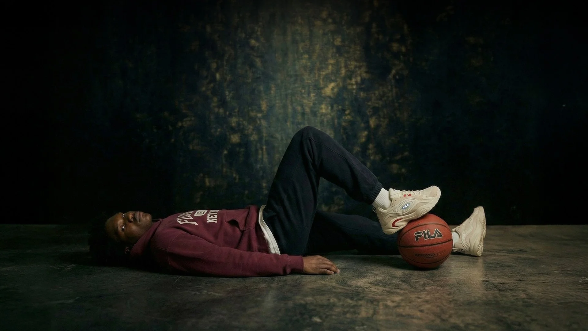 A person lying on the floor with a basketball under their leg, wearing a maroon hoodie, black pants, and white sneakers, looking at the camera.