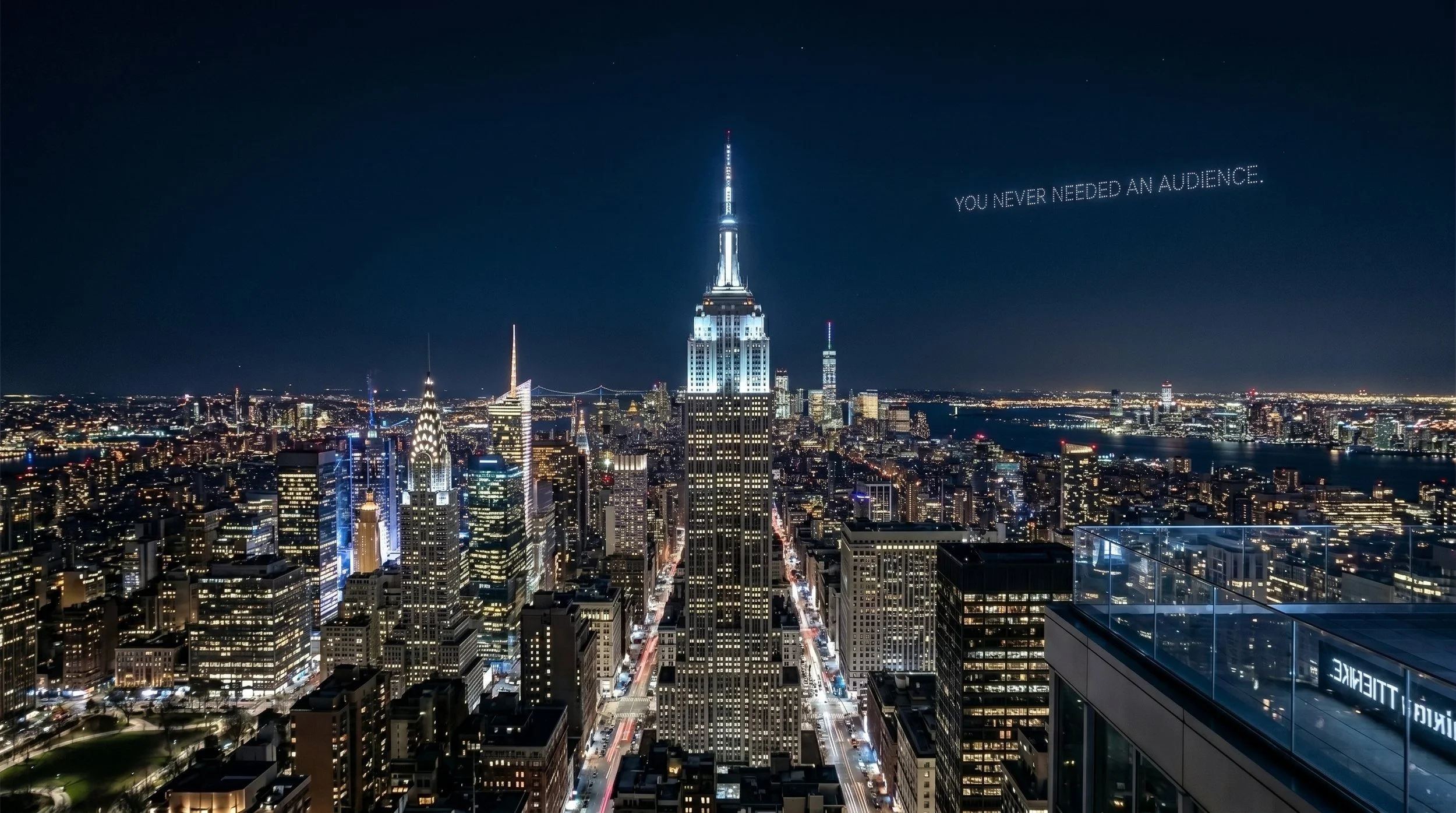 Nighttime aerial view of Manhattan skyline with illuminated buildings, including the Empire State Building, and the city lights reflecting on the water. The phrase "YOU NEVER NEEDED AN AUDIENCE" is written in the sky.