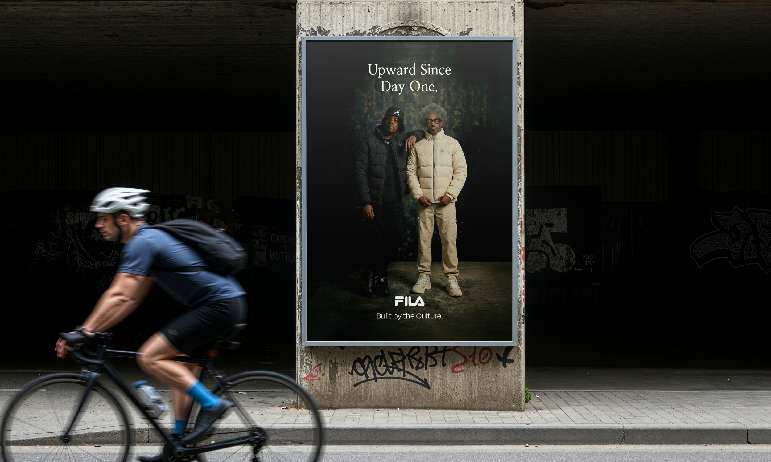 A cyclist riding past a poster on a city street. The poster features two men in jackets, and the text 'Upward Since Day One' with the FILA logo and the tagline 'Built by the Culture'.