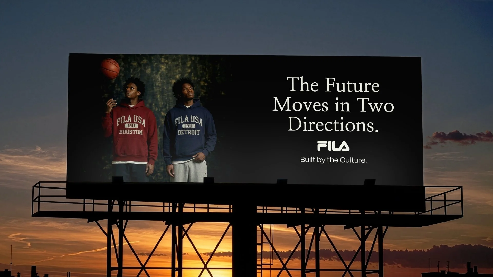 Billboard displaying two young men in hoodies with Fila USA logos, one in red and the other in blue, standing outdoors at sunset. The billboard has the text 'The Future Moves in Two Directions. FILA Built by the Culture.'