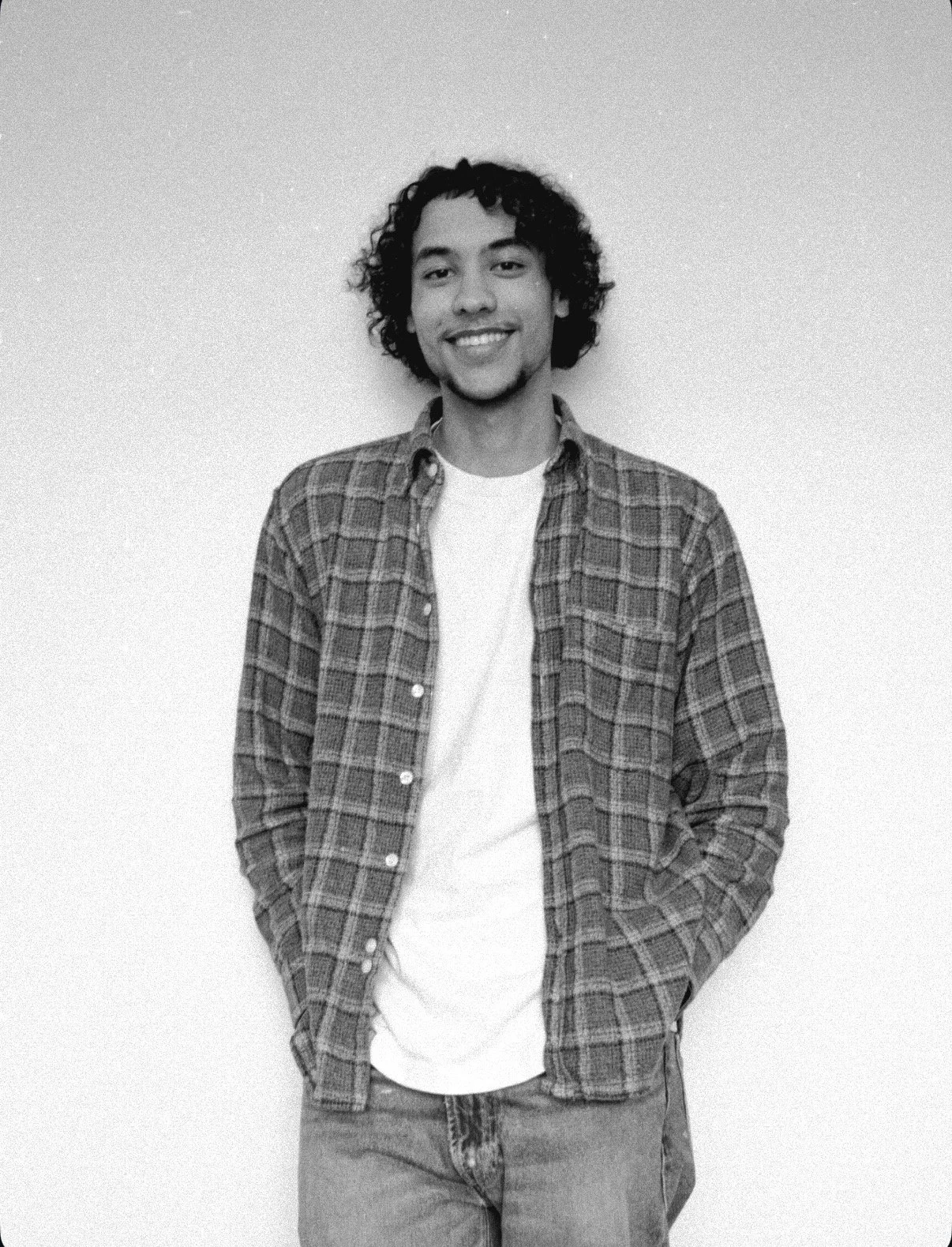 A young man with curly hair and a goatee smiling, wearing a checkered shirt over a white t-shirt, standing against a plain background.