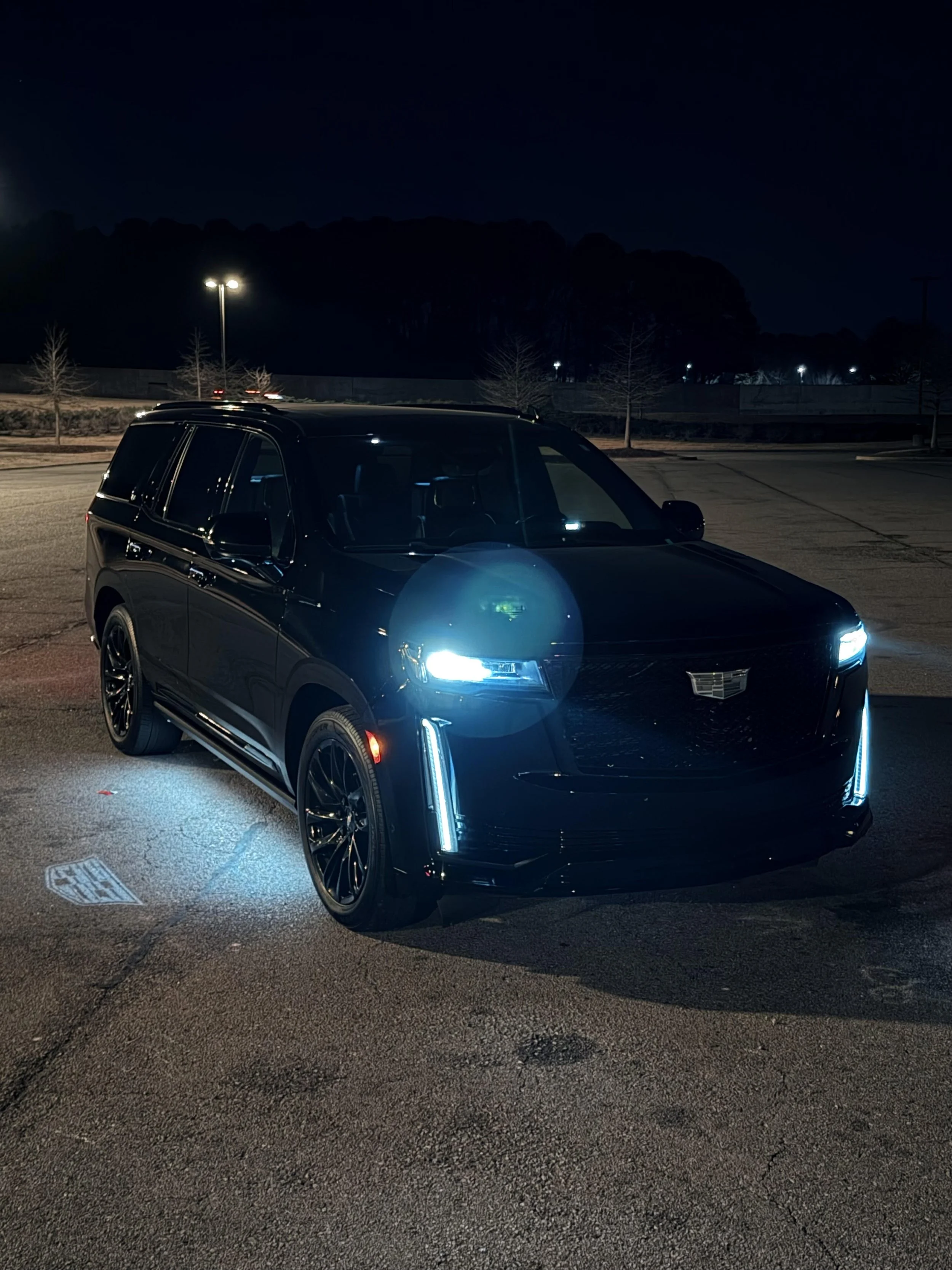 A black Cadillac SUV parked in a dimly lit parking lot at night, with its headlights and daytime running lights on. The background features streetlights, trees, and a dark hillside.