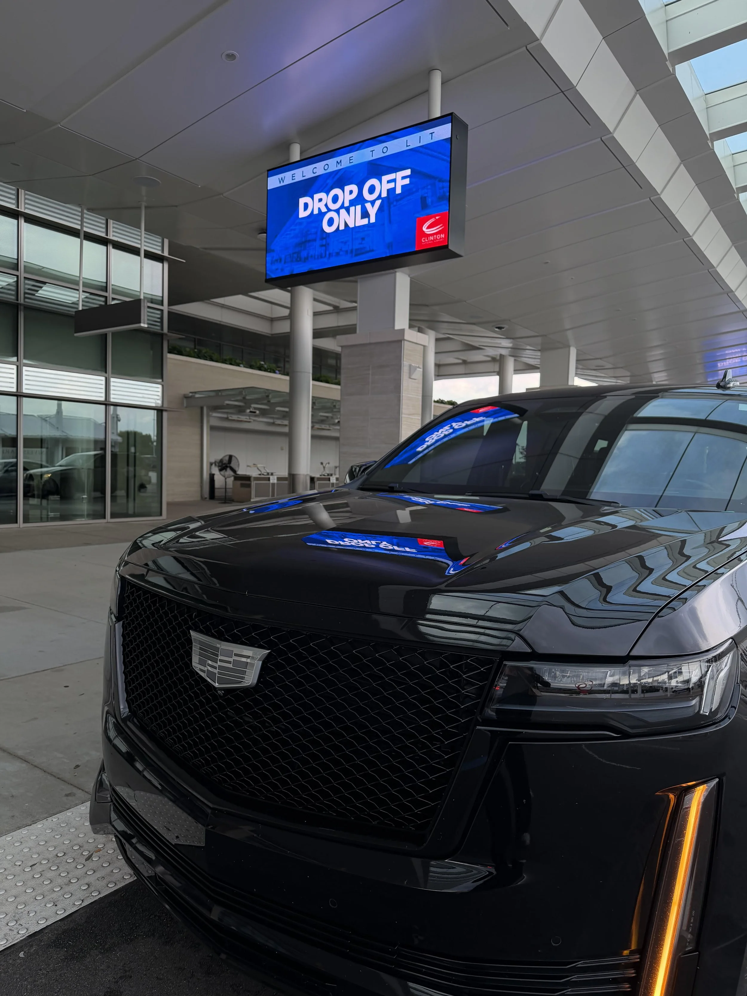 Black Cadillac parked at a curb in front of a modern building with glass windows. A sign overhead reads 'Drop Off Only' with the Clinton logo.