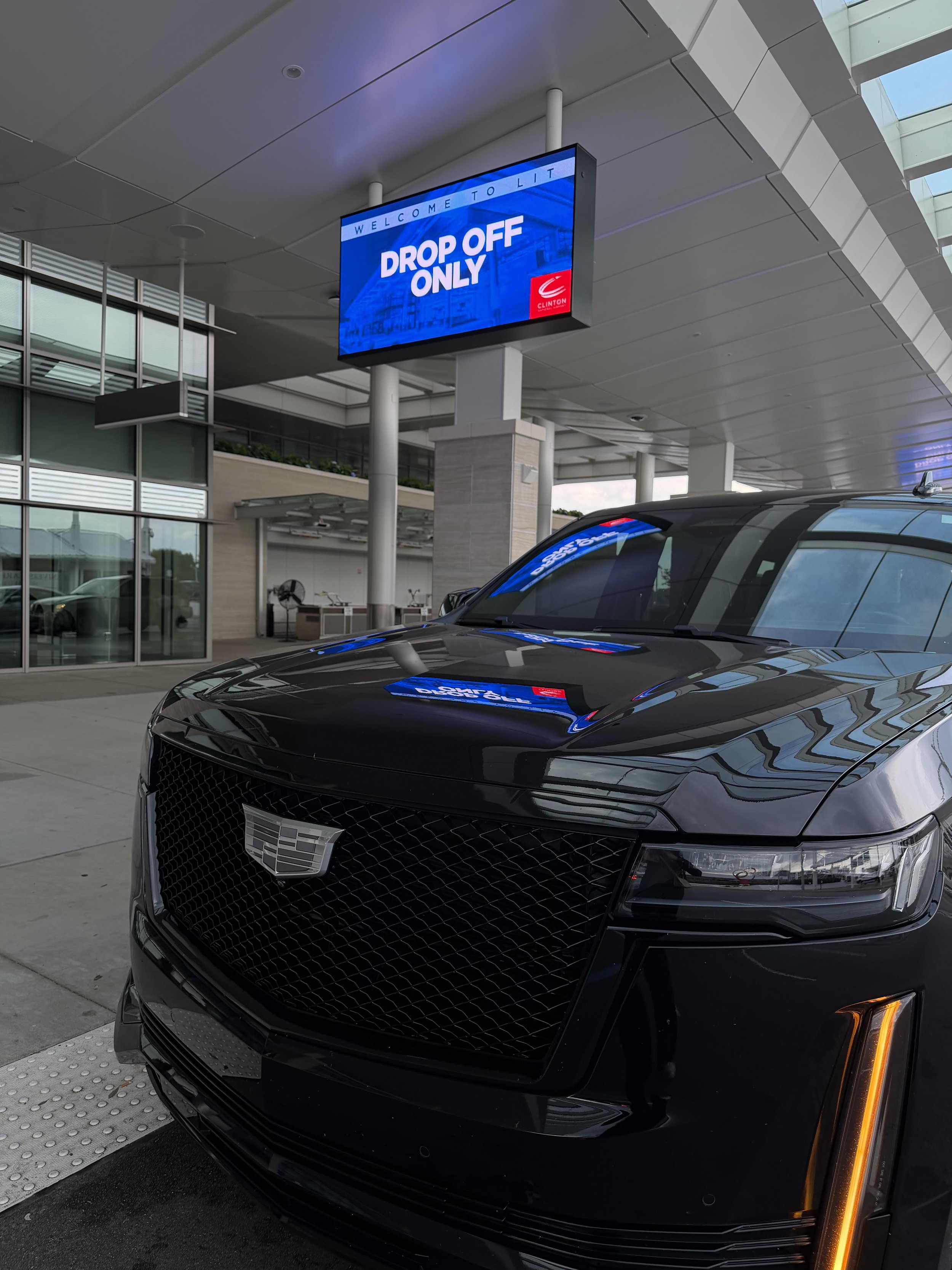 A black Cadillac car parked outside a modern building with glass windows and a digital sign that reads "Drop Off Only" with the Clinton logo.